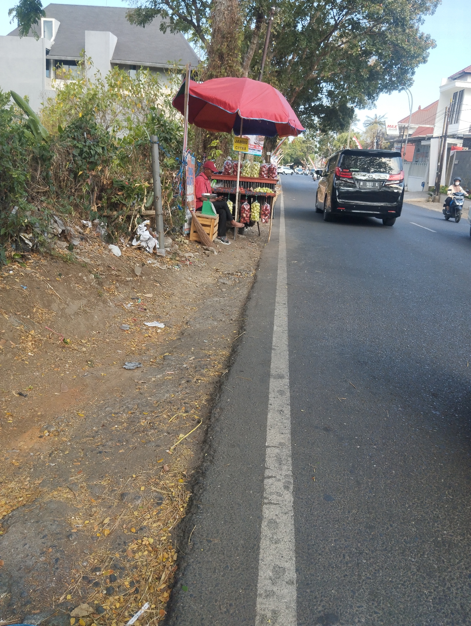 The image depicts a roadside scene in an urban environment, likely in Indonesia, with a fruit stand on the left and a street with traffic on the right. A red umbrella shades a small fruit stand, displaying various fruits such as apples and what appear to be guavas. A person wearing a red shirt and a red cap is seated behind the stand, possibly attending to customers or resting. A green scale is visible on the stand.
Adjacent to the fruit stand is a dirt and debris-filled embankment. The street itself is paved and has a white line marking the lane. A black minivan with license plate "N 1031 K" is driving in the right lane, and a person on a motorcycle is visible further down the road in the same lane. Buildings with distinct architecture, including a modern-style house with a grey roof, are visible on the right side of the street. Lush trees and foliage line the area, suggesting a moderately vegetated urban setting. The lighting indicates it is daytime, with sunlight casting shadows. The overall impression is a typical street view with local commerce and daily traffic.