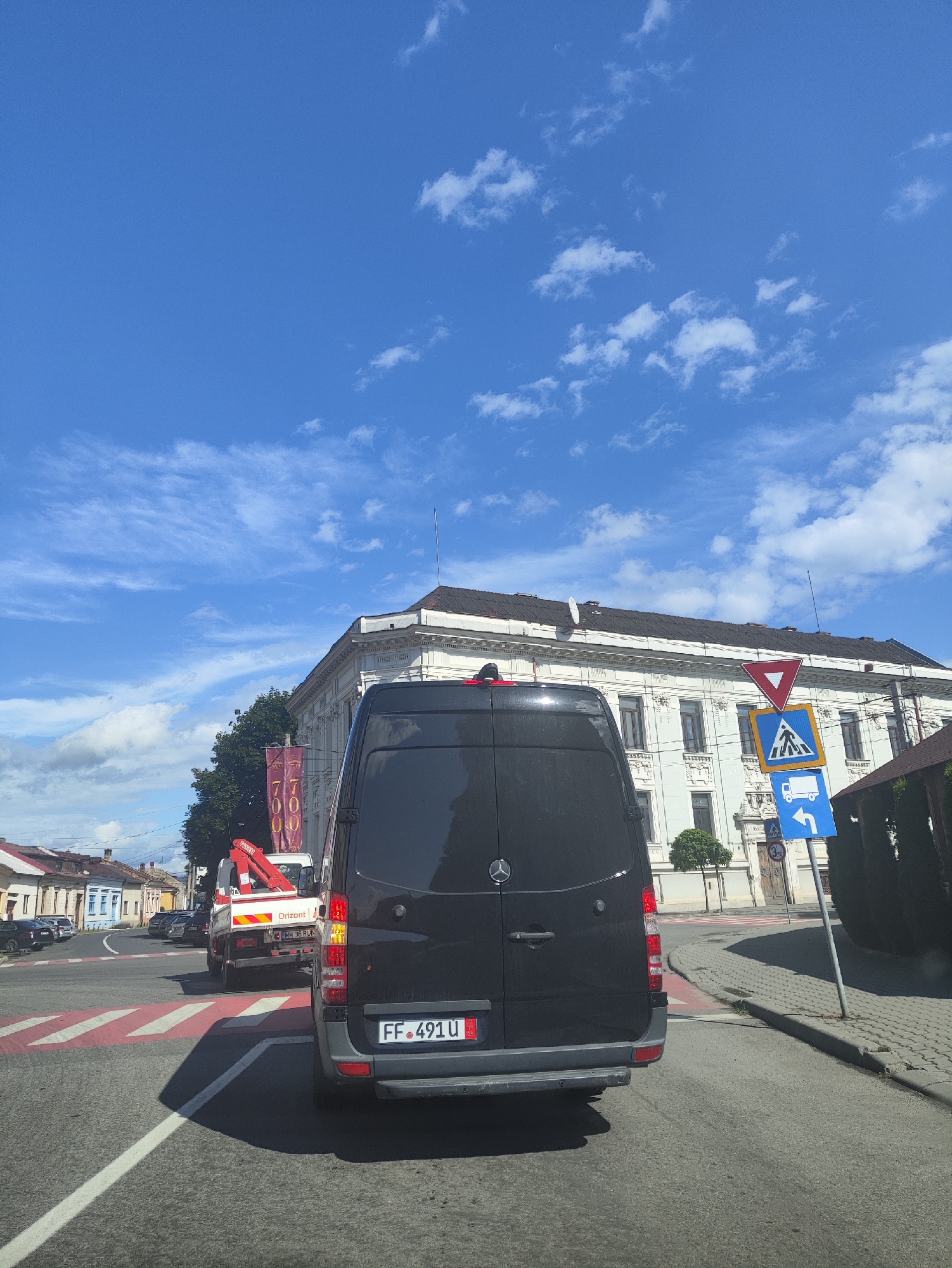 The image shows a view from a vehicle looking towards a street intersection in Sighetu Marmatiei, Romania. The sky is blue with scattered white clouds. In the foreground, the rear of a black Mercedes-Benz van is visible, with its license plate reading "FF 491 U". A red and white striped pedestrian crossing is marked on the asphalt.
Further ahead, a white truck with a red crane attachment is stopped at the intersection. The truck has text on its side that appears to read "Orizont". A banner with the text "7700" in red is hanging from a building on the right side of the street.
Dominating the background is a large, ornate white building with a dark roof. Several traffic signs are visible near this building. One is a red inverted triangle, a yield sign. Below it, a blue square sign with a white pedestrian symbol indicates a pedestrian crossing. Another blue sign below that shows a white truck with a curved arrow indicating a right turn. A speed limit sign indicating "3.5" is also visible.
To the left of the intersection, a row of colorful buildings with pitched roofs lines the street. Several parked cars are visible along this street. The scene depicts a typical street view in a Romanian town on a clear day.