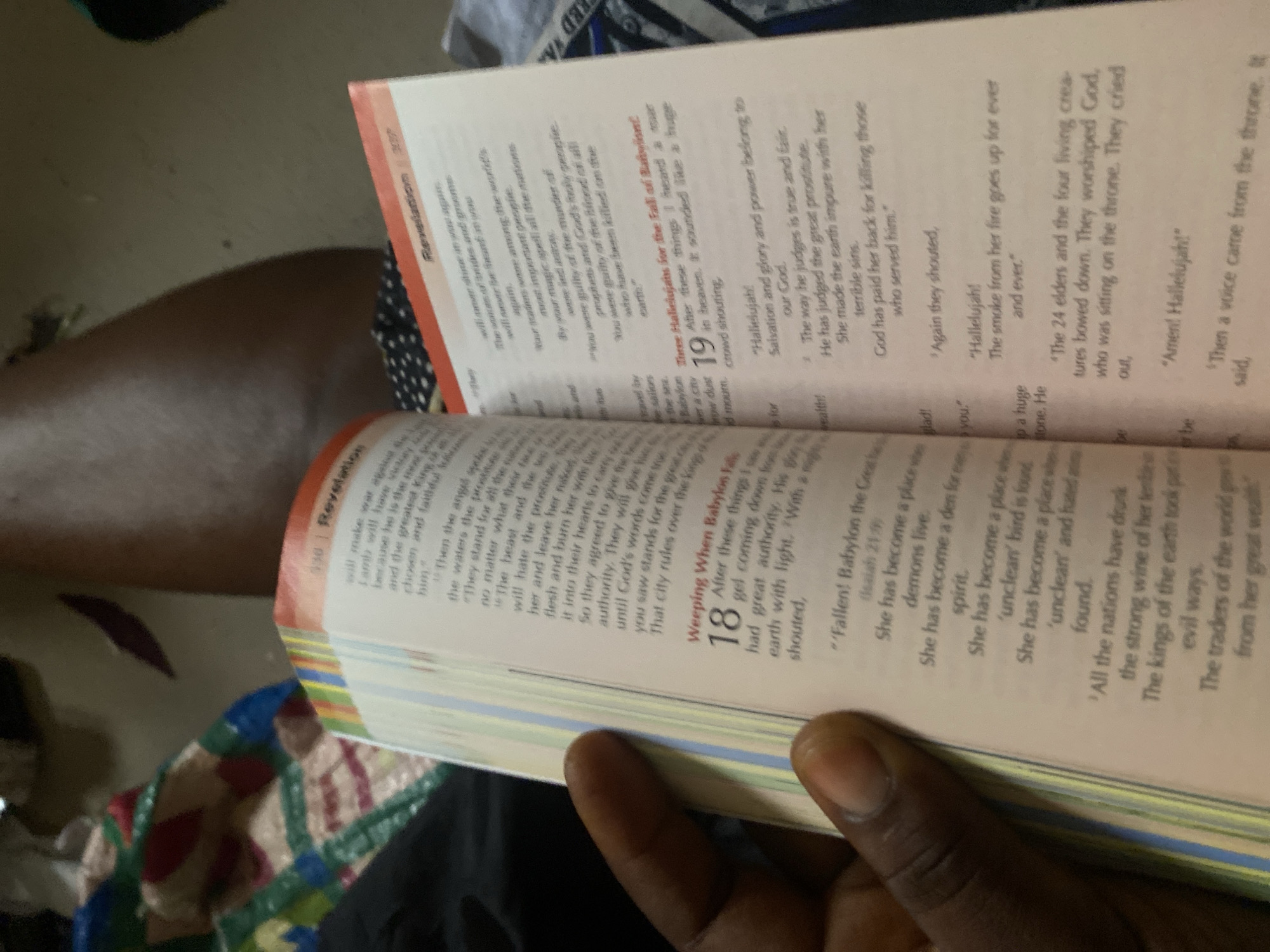 The image displays an open book, identified as the Bible, specifically showing passages from Revelation. A person's hand, with dark skin and a prominent thumb, is holding the book open, suggesting it is being read or examined. The pages are filled with text in two columns. On the left-hand page, the heading "Revelation" is visible at the top, followed by verses discussing "Weeping When Babylon Falls." A large numeral "18" marks the start of a new section. The right-hand page also features the heading "Revelation" at the top, followed by numbered verses, with "19" indicating "Three Hallelujahs for the Fall of Babylon." The lighting suggests an indoor setting, and the background is out of focus, showing what appears to be fabric with a colorful, patterned design. The image is a close-up of the book and hand, with no discernible elements of an outdoor environment or specific urban location. There is no information available in the image to determine its location in Jauro Yinu, Nigeria.