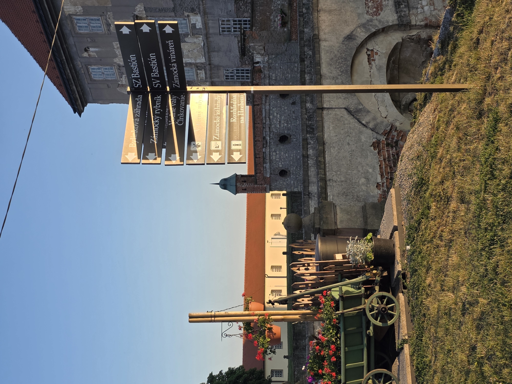 A close-up, eye-level, outdoor shot captures a signpost directing visitors to various attractions at the historical site of the town of Slavkov u Brna, Czech Republic, on a sunny day. The signpost is positioned to the right of the frame, with black directional signs pointing left and right, indicating destinations such as "Bylinková zahrada" (Herb Garden), "Zámocký rybník" (Castle Pond), "SZ Bastión," "SV Bastión," "Zámocká vináreň" (Castle Winery), and "Rozhledna na Hřebeni" (Lookout Tower on the Ridge).
In the foreground, a rustic green wooden cart is filled with colorful blooming flowers, including red geraniums and purple petunias. Beside the cart, a black pot contains a silvery-green plant. The cart and flowers are placed on a paved walkway with a grassy area beyond.
Behind the cart, a weathered wooden fence separates the walkway from a stone bridge with arched openings, revealing brickwork. Further back, a pale yellow building with a red-tiled roof stretches across the middle ground, its windows neatly aligned. A small, dark green tower with a pointed roof is visible above the stone structures.
To the right of the signpost, a section of an old, grey stone building with a red-tiled roof is visible. The building has multiple windows and shows signs of age with peeling plaster and visible brickwork. The overall scene suggests a historical and picturesque location, likely a castle or a fortified town. The lighting indicates it is either early morning or late afternoon, with the sun casting long shadows.