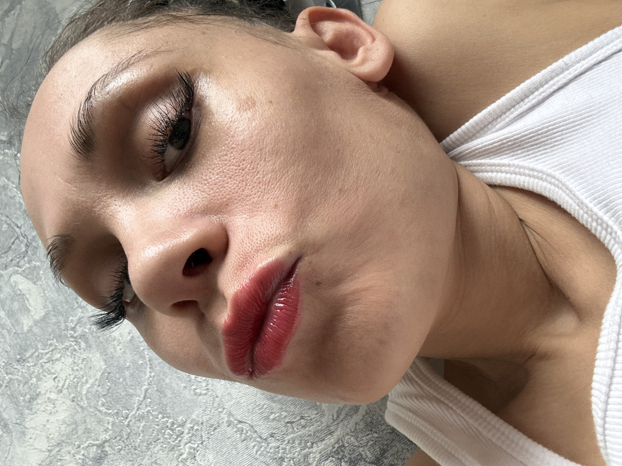 A close-up, angled portrait of a woman lying on a patterned surface. Her face is the primary focus, featuring prominent eyelashes, arched eyebrows, and glossy red lips. She has a fair complexion and her right ear is visible. She is wearing a white tank top. The photo does not provide any information to ascertain that it was taken in Rusanovo, Russia.