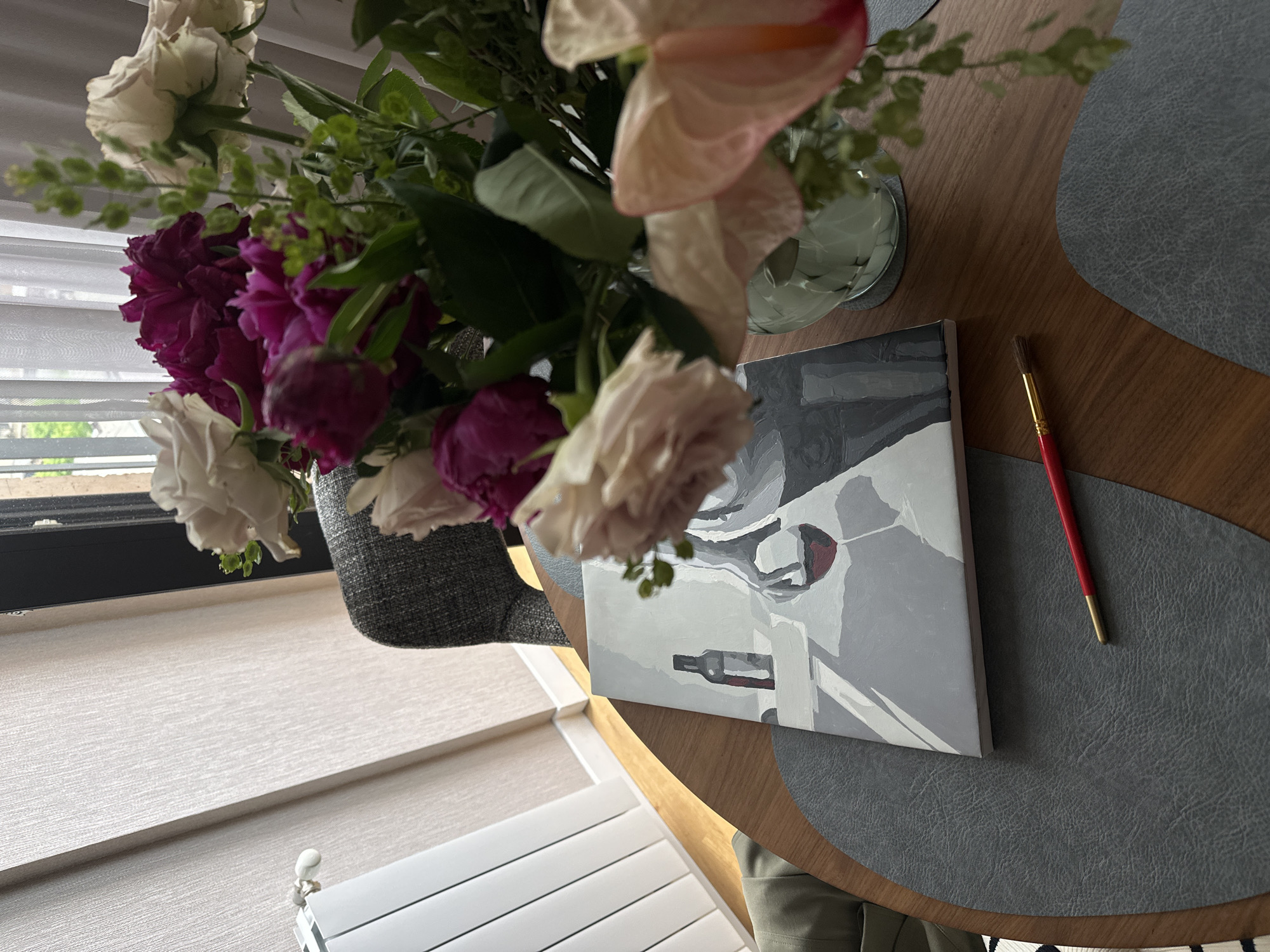 A vase of white and purple flowers sits on a round wooden table next to a painting of a still life and a paintbrush. The painting appears to depict a wine bottle and wine glass. The table is covered with a gray placemat. In the background, a window with closed white blinds can be seen. The image was taken from a slightly elevated perspective. The location is unknown and may not be in Yerevan, Armenia. No people are visible.
