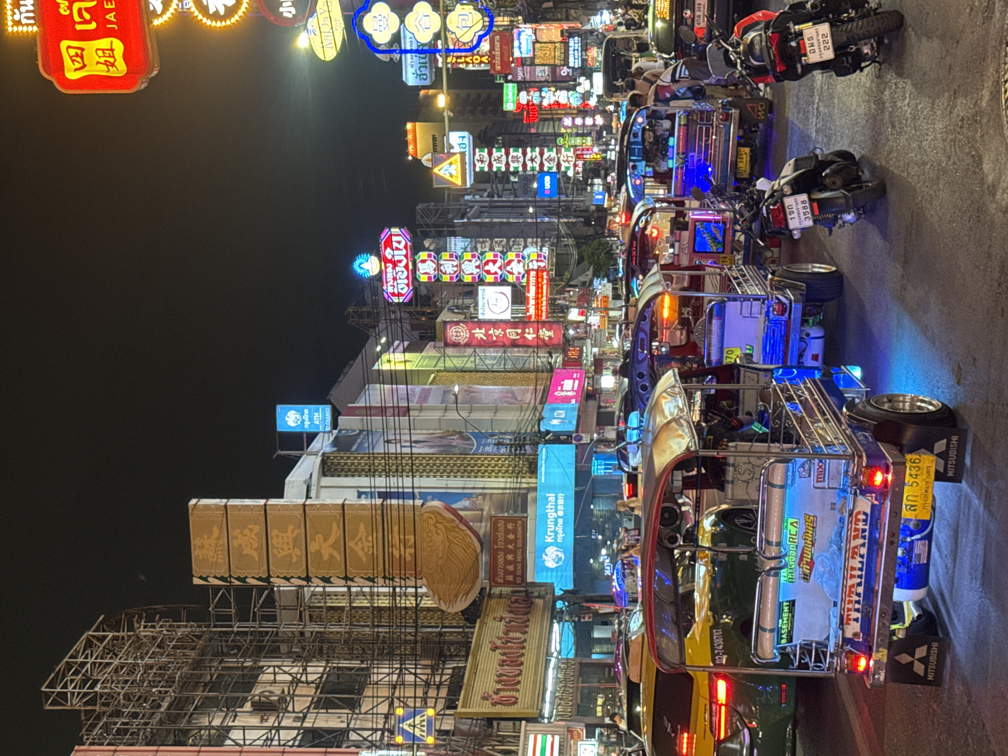 Here is a description of the image:
The image shows a busy street scene at night in Bangkok, Thailand. The street is lined with brightly lit signs in Thai and Chinese characters, many of them for gold shops and businesses. There is a row of "tuk-tuks", a type of three-wheeled motorized rickshaw, parked on the side of the road. The tuk-tuks are decorated with colorful lights and stickers. There are also motorcycles parked nearby. People are visible in the background, but they are not the main focus of the image. Overall, the image captures the vibrant and chaotic atmosphere of Bangkok at night.