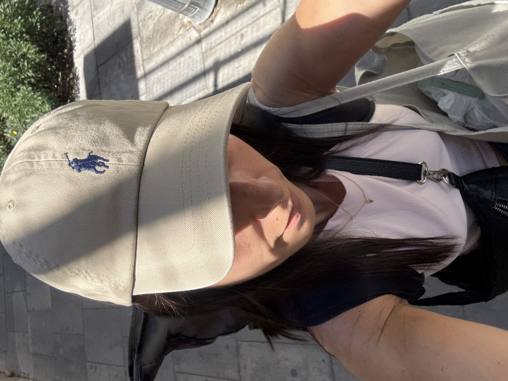 An overhead, rotated close-up shot captures a person, likely female, outdoors in Madrid, Spain, under bright sunlight. The subject is wearing a beige baseball cap with a dark blue embroidered Polo Ralph Lauren logo (a polo rider) clearly visible on the front. Her face is partially obscured by the cap and shadows, revealing her chin, mouth, and parts of her cheek. Long, dark hair cascades from beneath the cap.
She is dressed in a light pink or off-white ribbed top and wears a thin gold necklace with a small charm. A black strap, likely from a bag, crosses her chest. On her right side, she holds a light-colored tote bag, inside which a white plastic bag and other items are visible. Dark clothing, possibly a jacket or another layer, is seen around her shoulders.
The scene features a paved ground, resembling a sidewalk or pedestrian area with light-colored flagstones or concrete tiles, which is illuminated by strong sunlight, casting distinct shadows. Green foliage from bushes or small trees can be seen in the upper left background. The overall impression is a casual daytime moment in an urban setting.