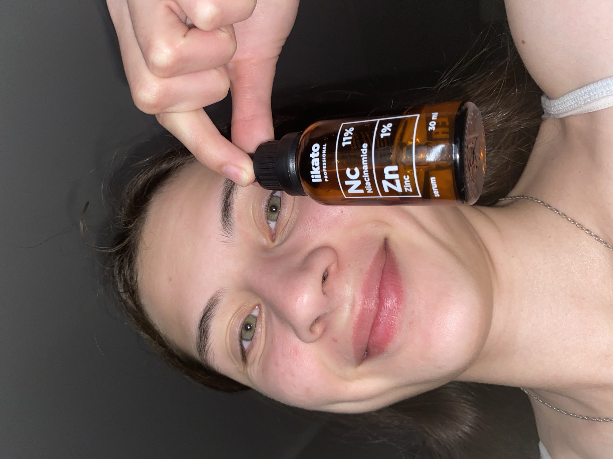 The image is a close-up portrait of a young woman, likely in her late teens or early twenties, holding a small brown glass bottle of Likato Professional Niacinamide and Zinc serum. The woman has fair skin with some redness on her cheeks, brown hair, and light green eyes. She is smiling and looking towards the camera. Her left hand is partially visible, holding the bottle near her face. The bottle has a black dropper cap and a label that reads "likato PROFESSIONAL", "Nc Niacinamide 11%", "Zn Zinc 1%", and "serum 30 ml". The background is dark and out of focus. There is no information available about the specific location within Votkinsk, Russia.