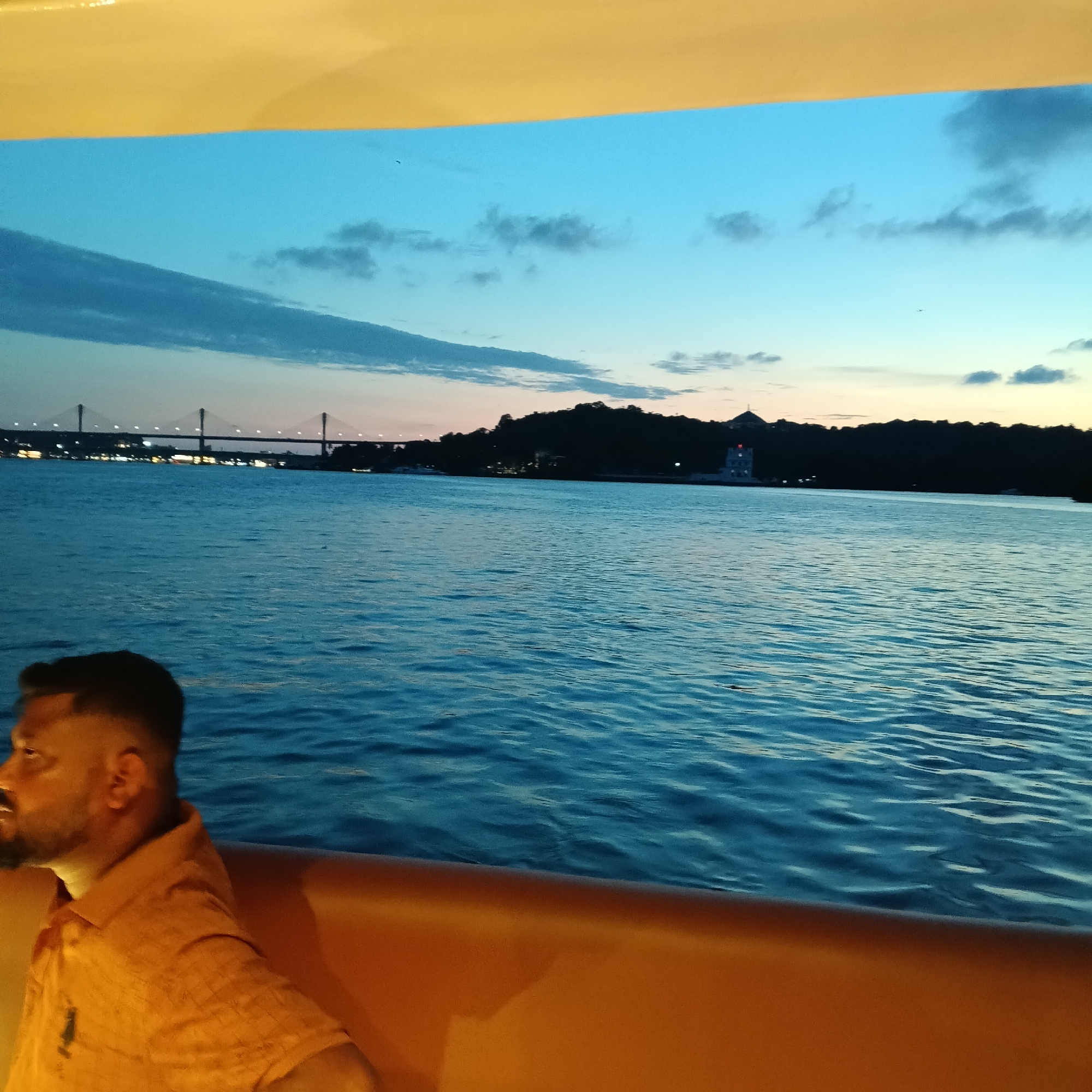 The image shows a person, likely male, sitting on a boat on a body of water, potentially the Mandovi River, in Britona, India. It is likely twilight or dusk based on the lighting. The person is wearing an orange shirt, and only his head and partial shoulder are visible. Behind the person is the river, which appears to be slightly choppy. Further in the background is the skyline of Panjim, featuring the Atal Setu bridge illuminated and connected to the city. An area of land covered in greenery and several buildings is visible on the right of the skyline. The sky has hues of blue and orange, indicating sunset or sunrise. The boat is likely illuminated with artificial lighting, creating warm tones on the person and the boat's interior.