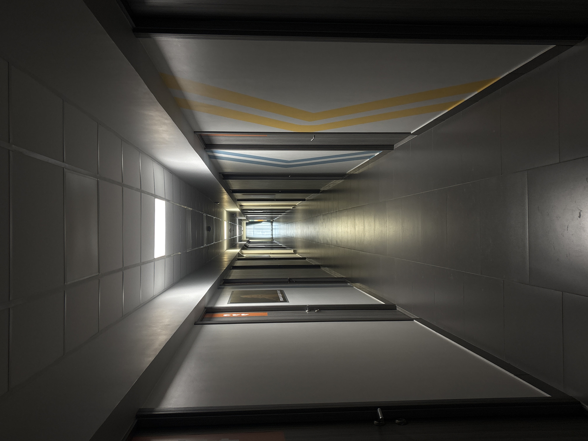 The image depicts a long, narrow corridor viewed from floor level in an office building in Perm, Russia. The walls are lined with doors, each marked with room numbers and signs. Fluorescent lighting fixtures illuminate the left side, while the right side is illuminated by an indistinct light source toward the distant end of the hall. Geometric patterns are painted on the ceiling in blue and yellow, forming a chevron design. The corridor walls are pale gray, with dark gray trim around the doors. The floor is dark. The perspective of the image creates an illusion of convergence towards the distant end. The scene appears unoccupied.
