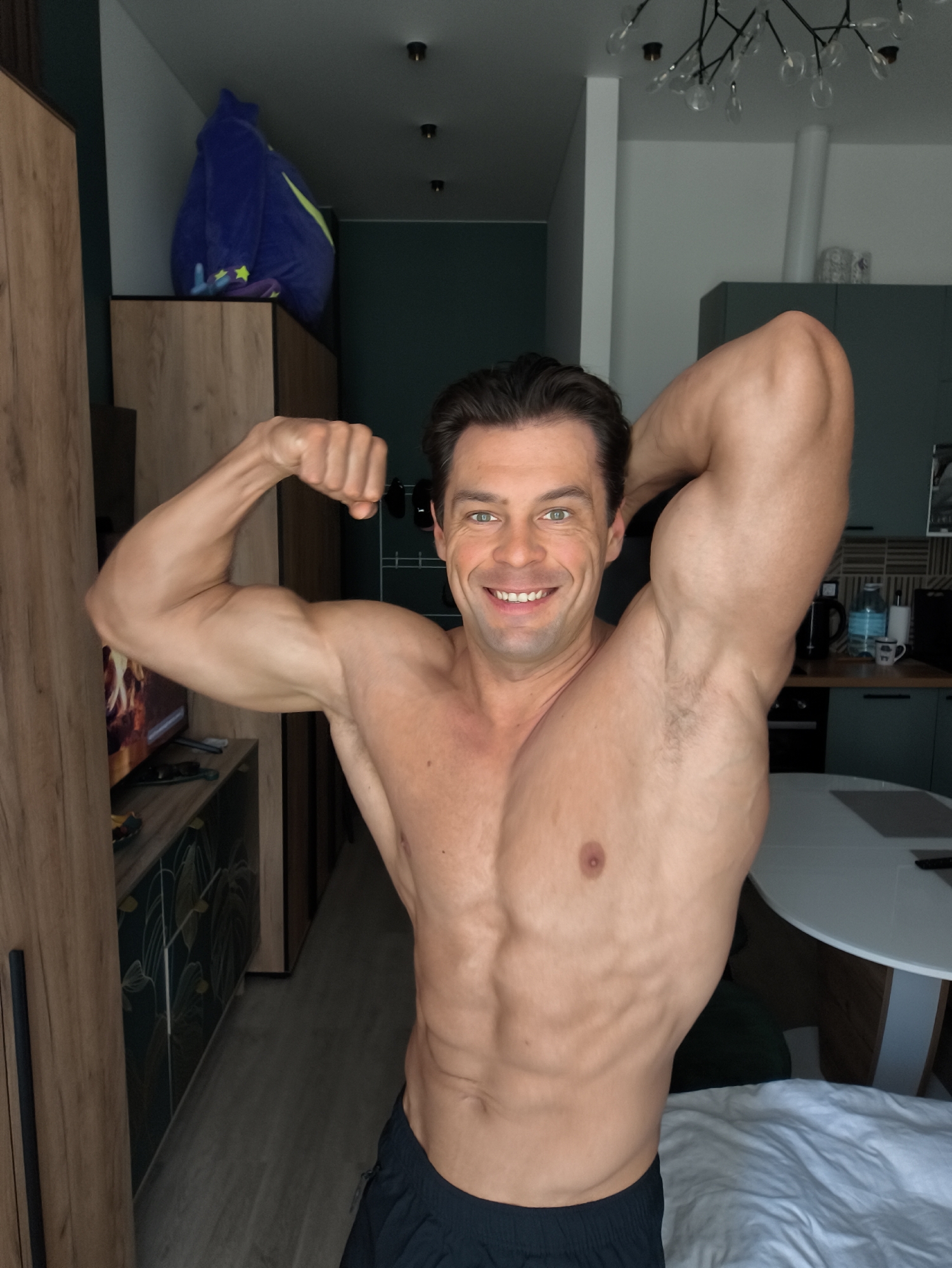 A muscular man with dark hair and green eyes is posing shirtless in a room, flexing his biceps. He is smiling and looking at the camera. The room appears to be a living space, possibly an apartment, with modern furniture and decor. In the background, there is a wooden wardrobe, a television, kitchen cabinets, and a round table with chairs. A white duvet covers a bed to the right of the frame. The lighting is natural, suggesting daytime. There are no identifiable landmarks or specific objects that would indicate the location as Moscow, Russia.