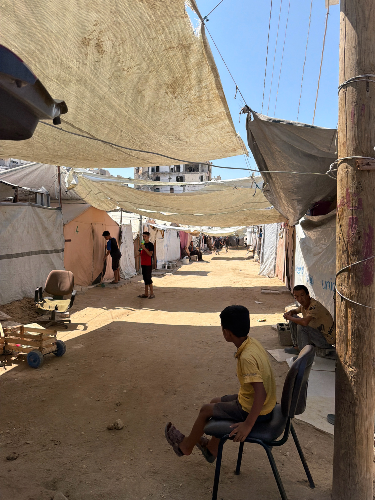 This photograph depicts a narrow, sun-drenched pathway in a refugee camp, likely located in the Gaza Strip, Palestinian Territories. Tents and makeshift shelters line both sides of the dirt path, covered by tarps and fabric awnings stretched overhead to provide shade. In the foreground, a young boy wearing a yellow t-shirt and dark shorts sits on a black office chair, facing away from the camera. To his right, a man in a light brown t-shirt and grey pants sits on a concrete block. Further down the path, two teenage boys are visible. One, in a dark blue shirt and black shorts, sweeps the ground with a broom. The other, in a red and black shirt and black pants, stands holding a green object, possibly a bottle. In the background, beyond the shelters, partially destroyed buildings suggest the impact of conflict. The overall environment is dusty and sparsely furnished, highlighting the austere conditions of the camp.