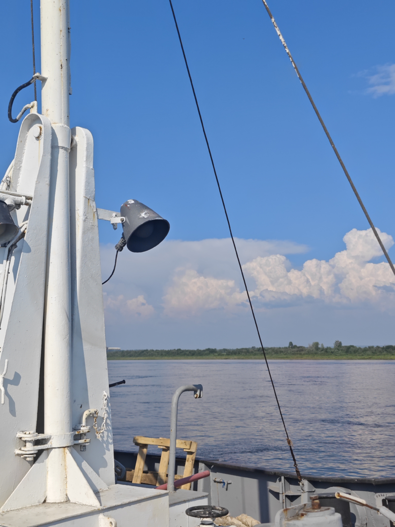 The image shows a view from a vessel on a body of water. In the foreground, a portion of the vessel's deck and superstructure is visible, primarily featuring a white metal mast assembly with attached fixtures, including a dark gray spotlight with a coiled cable. Another dark cable runs diagonally upwards across the frame, disappearing towards the top. The water is a deep blue with some ripples, and a distant tree-lined shore is visible on the horizon. The sky is a bright blue with scattered white cumulus clouds. The scene appears to be set during daylight with clear weather conditions. The location is identified as Verkhne-Pashino, Russia.