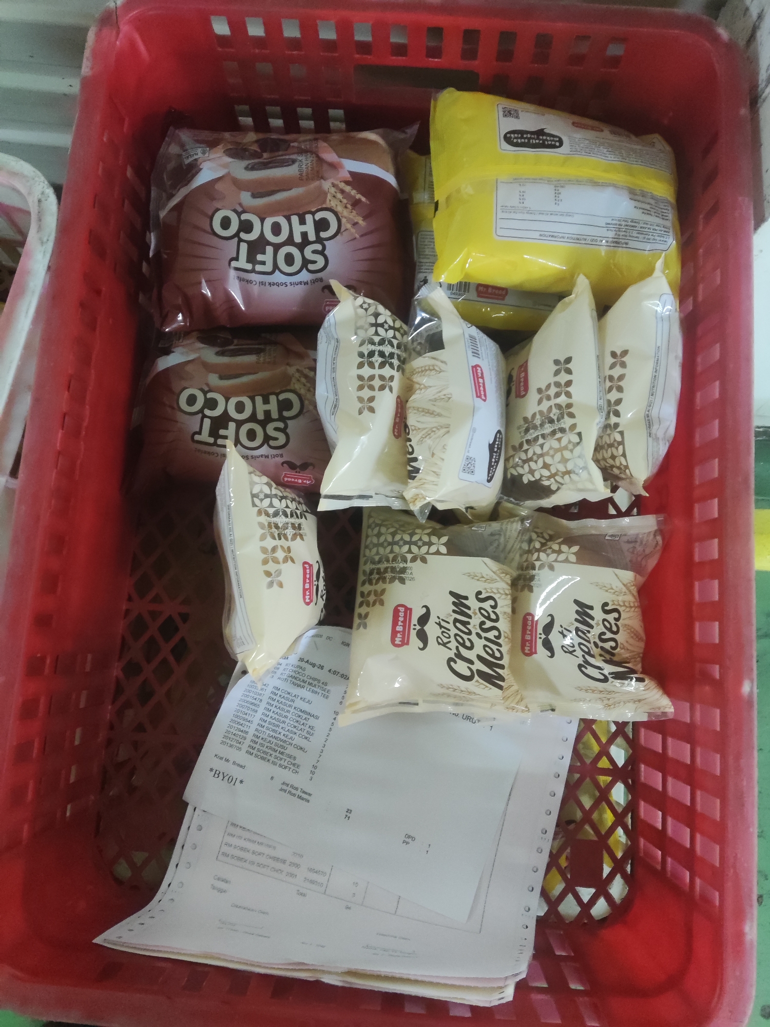 In a red plastic basket, a variety of food items are organized. The basket contains two large, yellow-wrapped packages, multiple individually wrapped packages of "Soft Choco" bread, and several packages labeled "Roti Cream Meises." The "Soft Choco" packages have a brown wrapper with text in white. The "Roti Cream Meises" packages are cream-colored with brown accents and feature the "Mr. Bread" logo and an image of a mustache. Beneath these food items, a stack of white paper documents is visible, some of which appear to be receipts or invoices with printed text and columns of numbers. The background is indistinct, but suggests an indoor environment. The scene is likely located in Trihanggo, Indonesia, given the presence of product names and potentially local documentation.