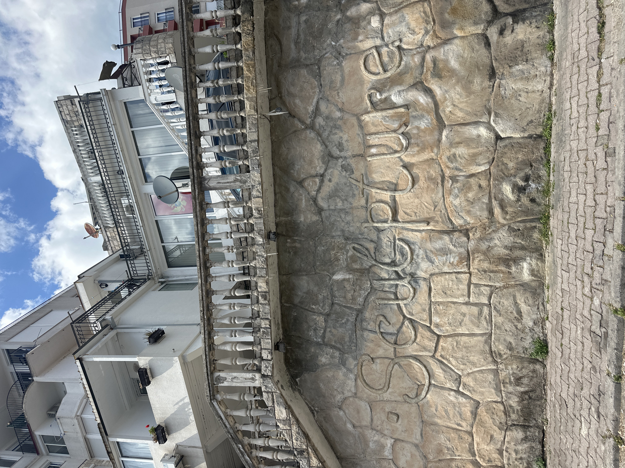 A medium-shot, eye-level perspective of a textured, stonework wall on a street in Budva, Montenegro, with a building extending to the left. The wall is predominantly beige with various shades of grey and brown, creating a rough, rocky texture. The word "Budva" is sculpted vertically on the wall, with each letter intricately embedded in the stonework. The wall stands alongside a grey, brick-paved sidewalk. To the left of the wall, a multi-story white building is visible, featuring balconies with white, ornate railings. Satellite dishes are affixed to the building's exterior. The sky is partly cloudy, visible above the building. The scene is set in daylight.
