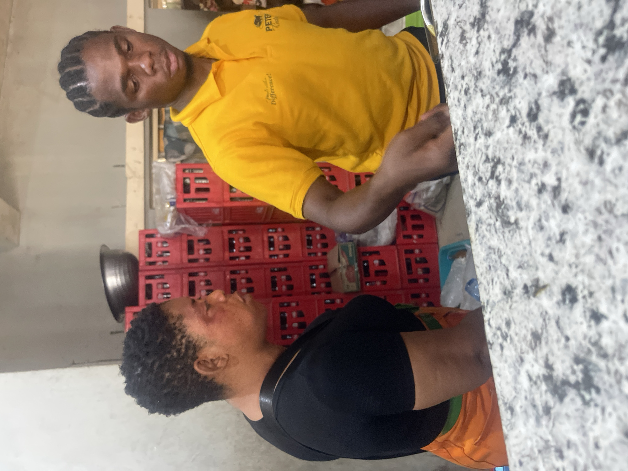 The provided image is oriented ninety degrees counter-clockwise, depicting an indoor commercial or storage setting in Zaria, Nigeria, featuring two individuals near a countertop with stacks of red beverage crates in the background.
When viewed upright, a young man with dark braided hair is prominently featured on the right side, wearing a bright yellow short-sleeved t-shirt. He looks down and slightly to his left with a neutral expression. The yellow shirt bears the text "PETER Cooler" (or "Cola") and "Make a Difference!", likely a brand or organizational motto. His hands are positioned close to a speckled gray and white countertop.
On the left side of the frame, another person, possibly a woman, with short, dark, curly hair, is visible from the side. They wear a black top and an orange lower garment, and are looking downwards, engaged with something on or below the countertop.
The background is dominated by numerous stacks of red plastic crates, commonly used for beverage storage or transport, reinforcing the commercial nature of the location. A dark metal bowl is also visible on a shelf behind the second individual. The lighting suggests an indoor daytime setting or a well-lit interior, indicating an active work environment.