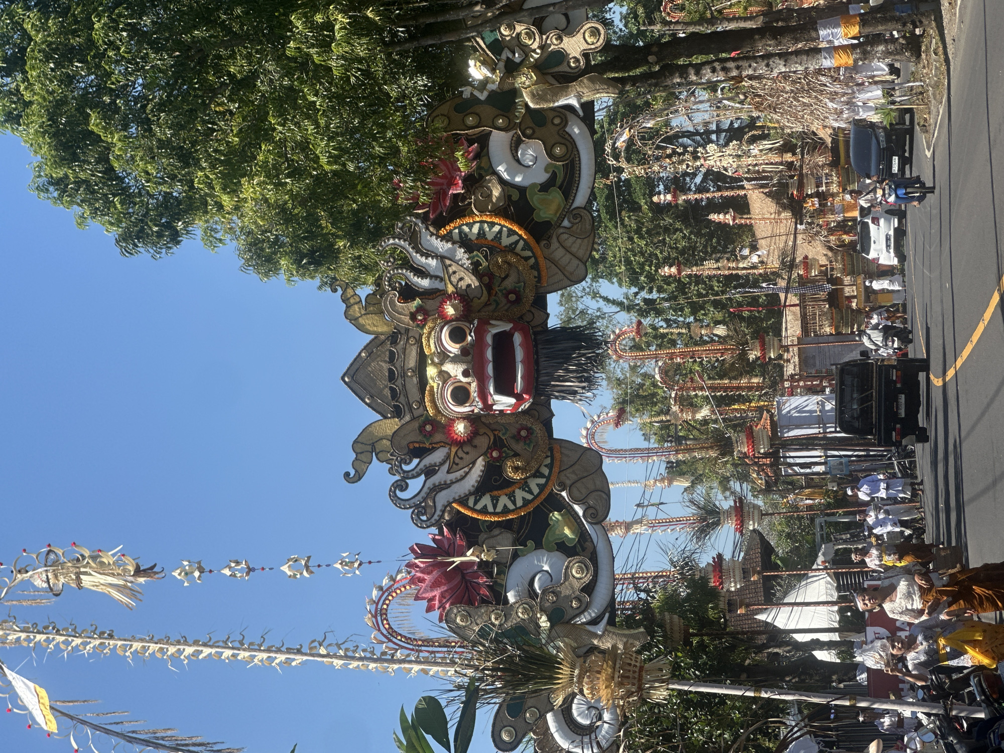 The image depicts a street scene in Sukawati, Indonesia, during a festival or procession. Dominating the foreground is an elaborate archway adorned with a Balinese Barong mask, intricate gold and natural fiber decorations, and floral elements. The arch spans across the road, which is paved and has a yellow line marking.
On the street, several people are visible, some in traditional Balinese attire, including women in sarongs and ornate blouses, and men in white robes. Some are walking, while others are on motorcycles or in cars. A black pickup truck is in the left lane, and other vehicles, including a white sedan and a motorcycle, are further down the road.
The sides of the street are lined with trees and a variety of decorative structures, characteristic of Balinese celebrations. Tall, elaborately decorated poles topped with woven straw and red accents are spaced along the road. Some buildings with thatched roofs are visible in the background. The sky is clear and blue. The overall impression is of a vibrant cultural event taking place in a tropical setting.