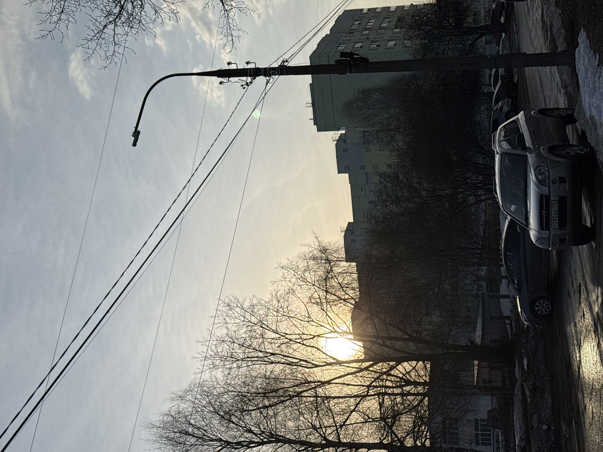 The photograph depicts a street scene in Chaikovsky, Russia. Bare trees dominate the foreground, obscuring the setting sun. In the background are apartment buildings. Utility poles and power lines are visible. Several parked cars line the right side of the street, along with snow and ice. The license plate of the vehicle closest to the camera is visible, T785AA159.