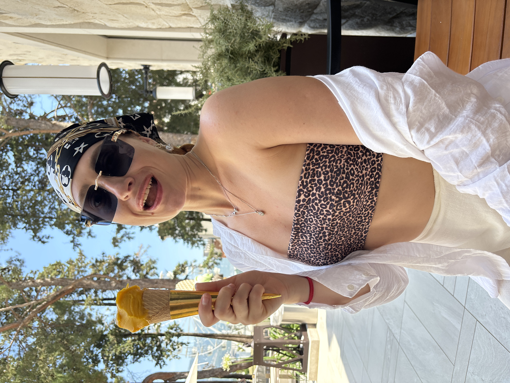 In Budva, Montenegro, a woman with blonde hair wears a black and white patterned headscarf, large dark sunglasses, and a leopard print bandeau top, with a white unbuttoned shirt draped over her shoulders. She is holding a cone of yellow ice cream in her right hand, and her mouth is open in an excited expression. She wears a silver necklace. The background features trees and a stone building with outdoor lighting fixtures. The scene appears to be outdoors during daylight.