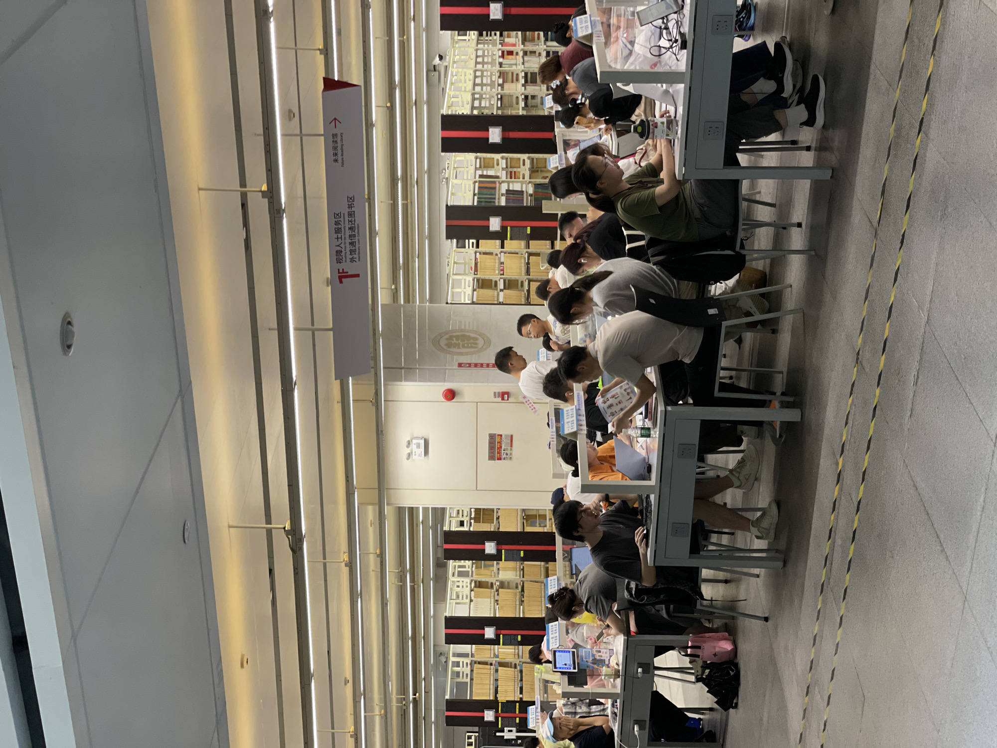 In a well-lit library setting in the Tianhe District of China, rows of individuals are seated at desks, deeply engrossed in their studies. Bookshelves filled with reading materials line the walls in the background, suggesting a place of learning and research. Fluorescent lights illuminate the space. The individuals are engaged in various activities such as reading, writing, or working on their laptops. Their postures and expressions indicate concentration and focus.
