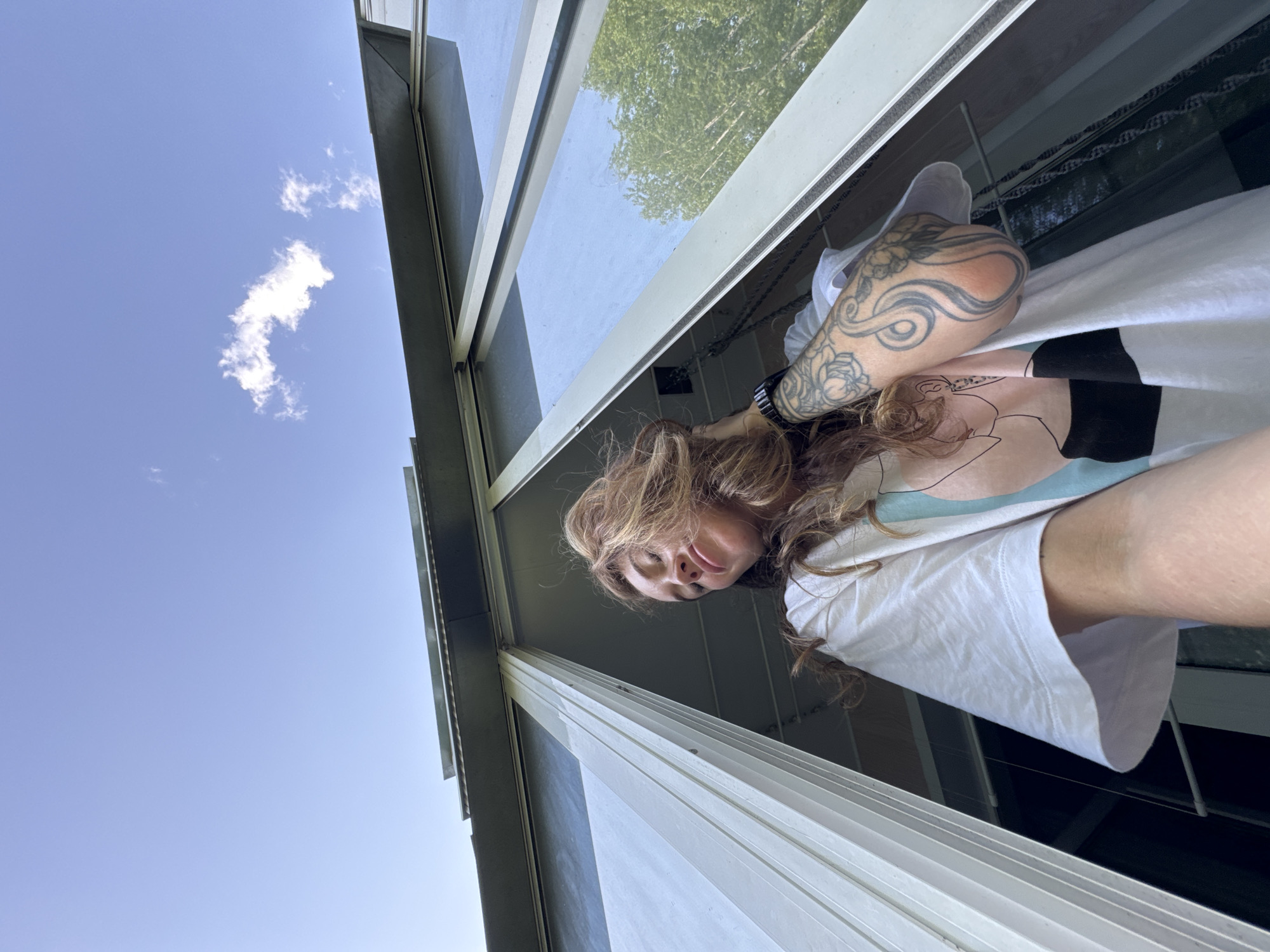 A low-angle shot looking up features a woman positioned near a window. She has wavy brown hair and is wearing a white t-shirt with a black and pastel design. Her left arm is visible, displaying tattoo art.
The view through the window includes a bright blue sky with a single cumulus cloud. The window frame is visible, with a backdrop of a green tree visible through the window glass. The location is in the city of Chaikovsky, Russia.