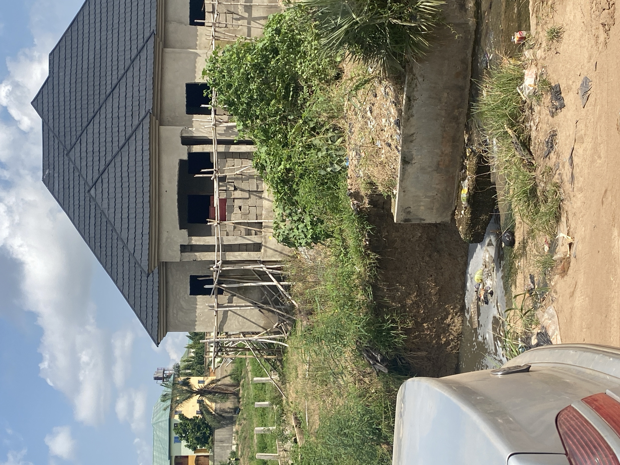 This image captures an outdoor scene in Ado, Nigeria, likely a residential or developing area. In the foreground, the rear left quarter of a silver car is visible. Beyond the car, a partially constructed building with a dark tiled roof and concrete block walls stands. Wooden scaffolding supports the construction. Overgrown vegetation, including a large bush, surrounds the building and the construction site. In the mid-ground, a dirty, shallow body of water with visible debris, including plastic bags and bottles, flows. Concrete structures are present along the water's edge. In the background, other residential buildings with colorful facades and tiled roofs are visible, along with palm trees and a water tower. The sky is partly cloudy.