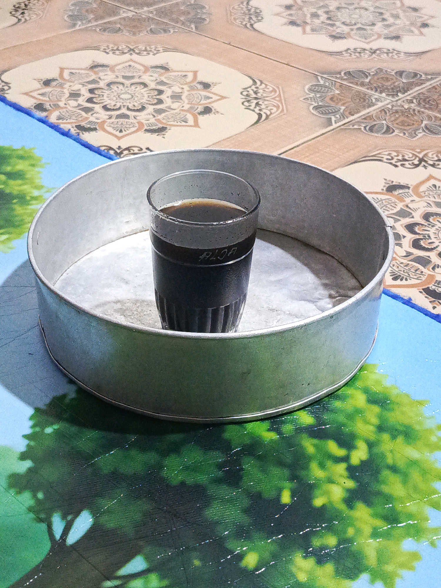 The image shows a glass of dark liquid, likely coffee or tea, placed inside a circular metal pan. The pan sits on a blue mat with a green, stylized tree pattern. In the background, a patterned tile floor is visible. The lighting suggests an indoor setting, possibly a home. The location is identified as Megang Sakti I, Indonesia. No people are present in the image.