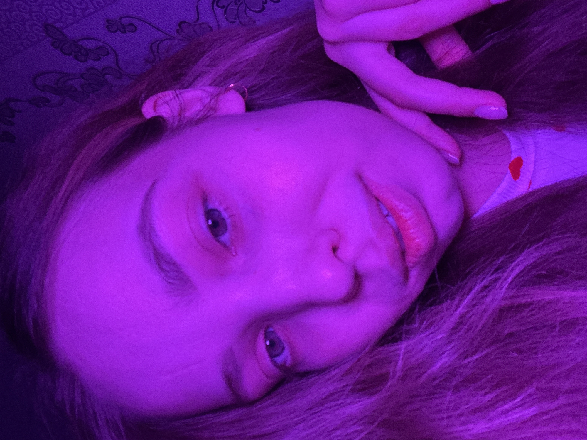 The image depicts a close-up, head-and-shoulders shot of a young individual, likely female, illuminated by strong magenta/purple lighting. The subject faces the camera directly with a slight upward tilt. Their right hand is positioned beneath their chin, with fingers slightly curled and visible, exhibiting light-colored nail polish. Long, light-colored hair, tinted purple by the lighting, frames the face and falls over the shoulders. The individual has light-colored eyes, which appear blue or green under the purple cast, and their lips are slightly parted, appearing glossy. A small hoop earring is visible in the right ear. Part of a white garment with a small red heart emblem is discernible around the neck and chest area. The background consists of a wall covered with patterned wallpaper featuring dark floral or vine-like motifs against a subtly lighter base, consistent with an indoor domestic setting. The specific location within the city of Chaikovsky, Russia, cannot be determined from the visual evidence provided.