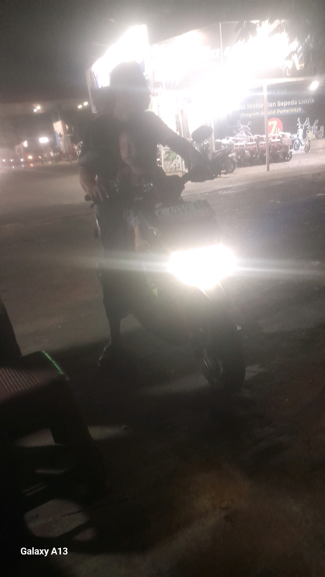 The image depicts a nighttime scene at what appears to be a government subsidy distribution point for electric motorcycles in Jekulo, Indonesia. The foreground is dominated by a motorcycle with its bright headlight illuminating the surroundings, obscuring much of the details. A person is seated on the motorcycle, with what appears to be another person behind them, though their features are indistinct due to the lighting and the quality of the image.
In the background, a brightly lit structure can be seen, with signage that reads "Motor Dan Sepeda Listrik Program Subsidi Pemerintah," translating to "Electric Motorcycles and Bicycles Government Subsidy Program." This suggests a government initiative aimed at promoting electric transportation. Multiple motorcycles are parked in neat rows behind the main subject, some with delivery boxes on their rear racks, indicating they may be intended for commercial use or distribution. The overall environment suggests an organized event or program taking place in an outdoor setting at night. The presence of the "Galaxy A13" watermark in the bottom left corner indicates the image was captured with a Samsung Galaxy A13 mobile phone.
