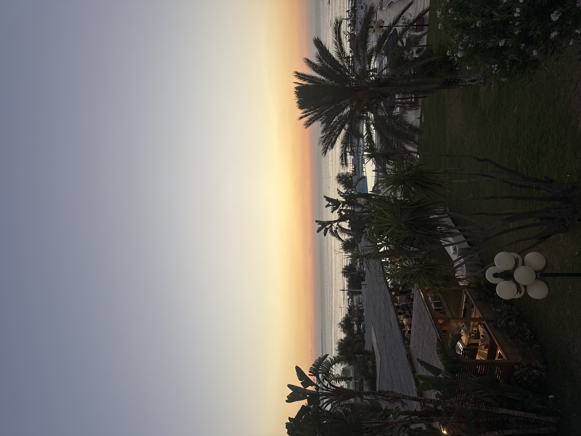 The image depicts a view of the sea and a coastal resort area at dusk. In the foreground, there is lush green grass and palm trees. To the left, a thatched-roof structure with outdoor seating and food preparation areas is visible, with lights illuminating the interior and tables. A multi-bulb lamppost stands on the grass in the lower right quadrant. Beyond the resort area, a swimming pool is visible, surrounded by lounge chairs and umbrellas. The sea stretches out to the horizon, with a faint glow from the setting sun casting warm hues of orange and yellow across the sky, which transitions to a soft blue above. The location is identified as Isola delle Femmine, Italy.
