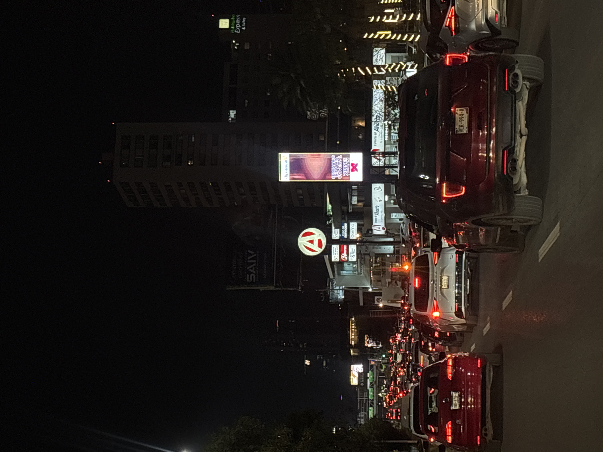 The image shows a nighttime street scene in San Bernardino Tlaxcalancingo, Mexico, with heavy traffic. Car tail lights illuminate the road. Tall buildings and commercial signs, including one for "Farmacias de Ahorro," SAIV, and a billboard, are visible in the background.