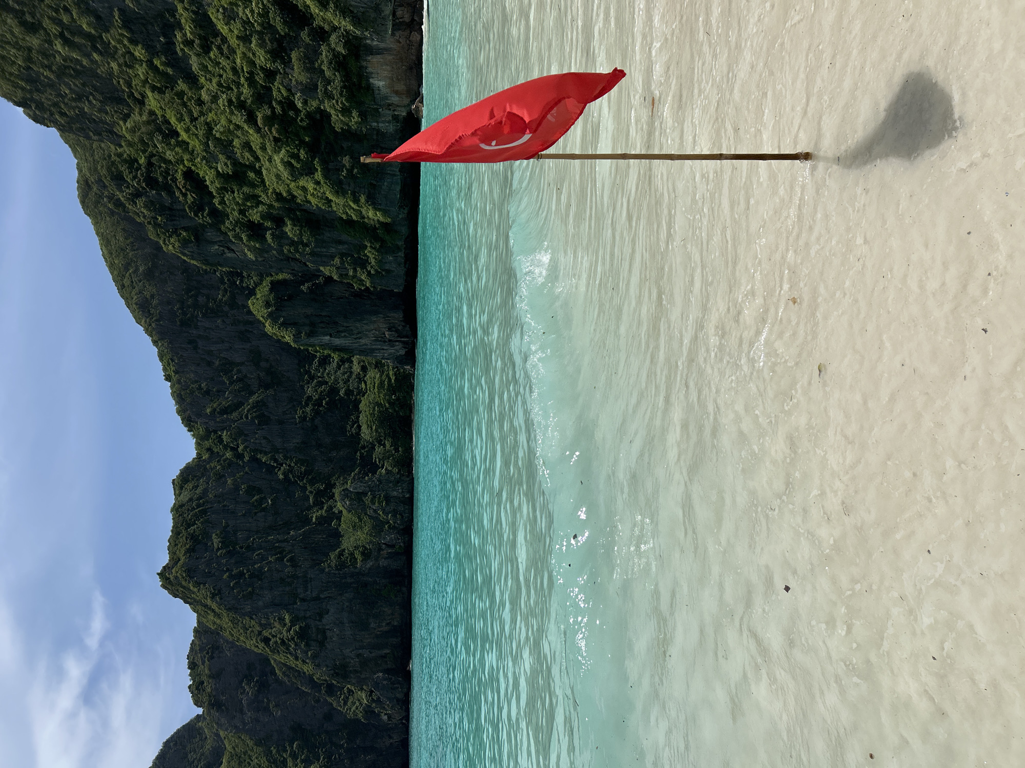 A red flag with a white crescent moon and star, the flag of Turkey, is planted in the shallow, clear turquoise water of a tropical beach. The flag is attached to a wooden pole, which is partially submerged in the sand. Gentle waves lap at the shore, creating a shimmering effect on the water's surface. In the background, lush green, rocky cliffs rise steeply from the water's edge, covered in dense vegetation. The sky above is a clear blue with a few wispy white clouds. The scene suggests a serene and beautiful natural landscape, possibly a secluded cove or bay. The location is identified as Ao Nang, Thailand.