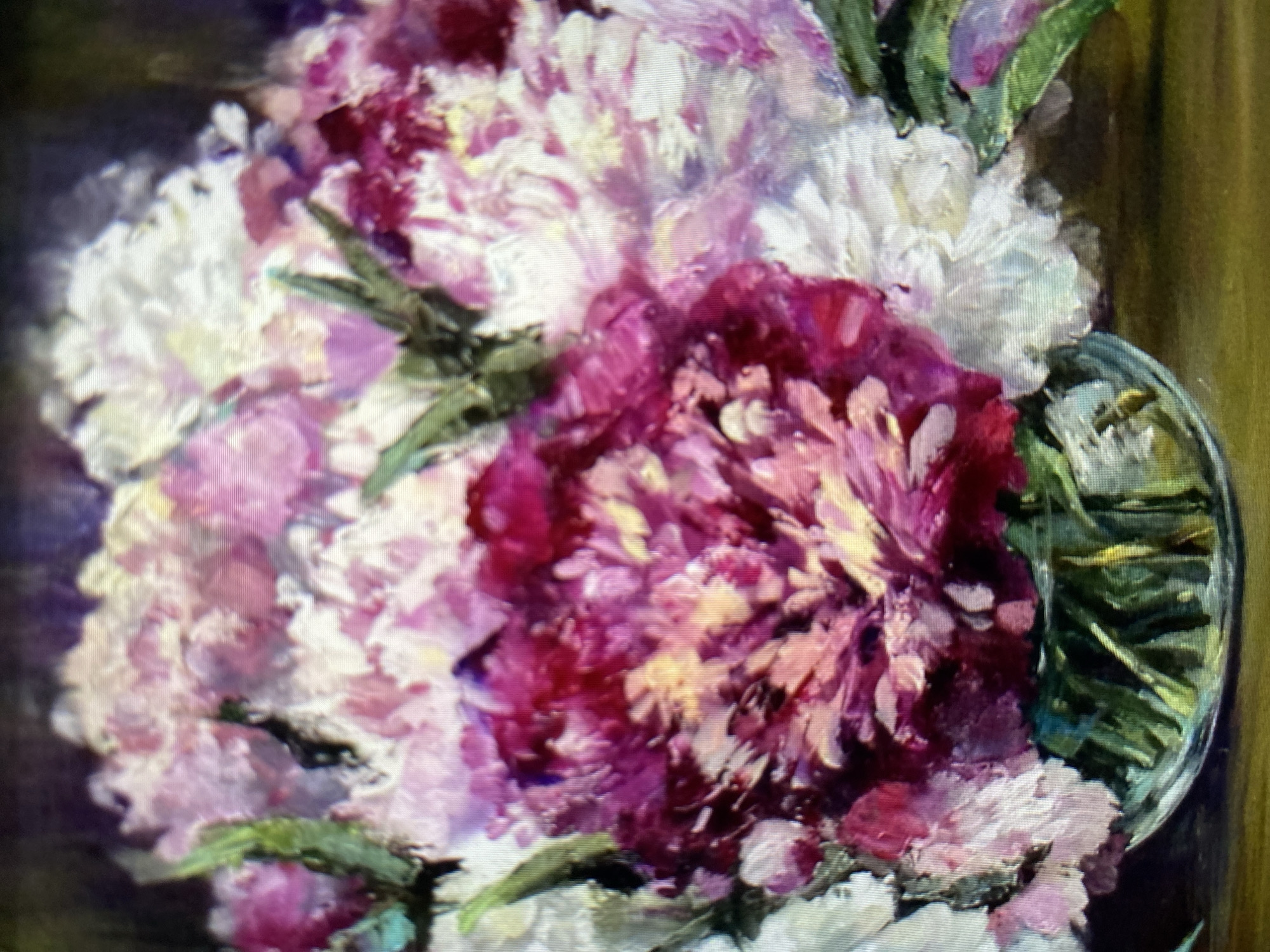 A close-up oil painting depicts a bouquet of peonies in a glass vase. The peonies are a mix of deep pink, light pink, and white, with visible brushstrokes adding texture and depth. Green stems are visible in the clear glass vase, which rests on a brown surface. The background is dark and blurred, focusing attention on the vibrant flowers. The painting appears to be a still life composition.