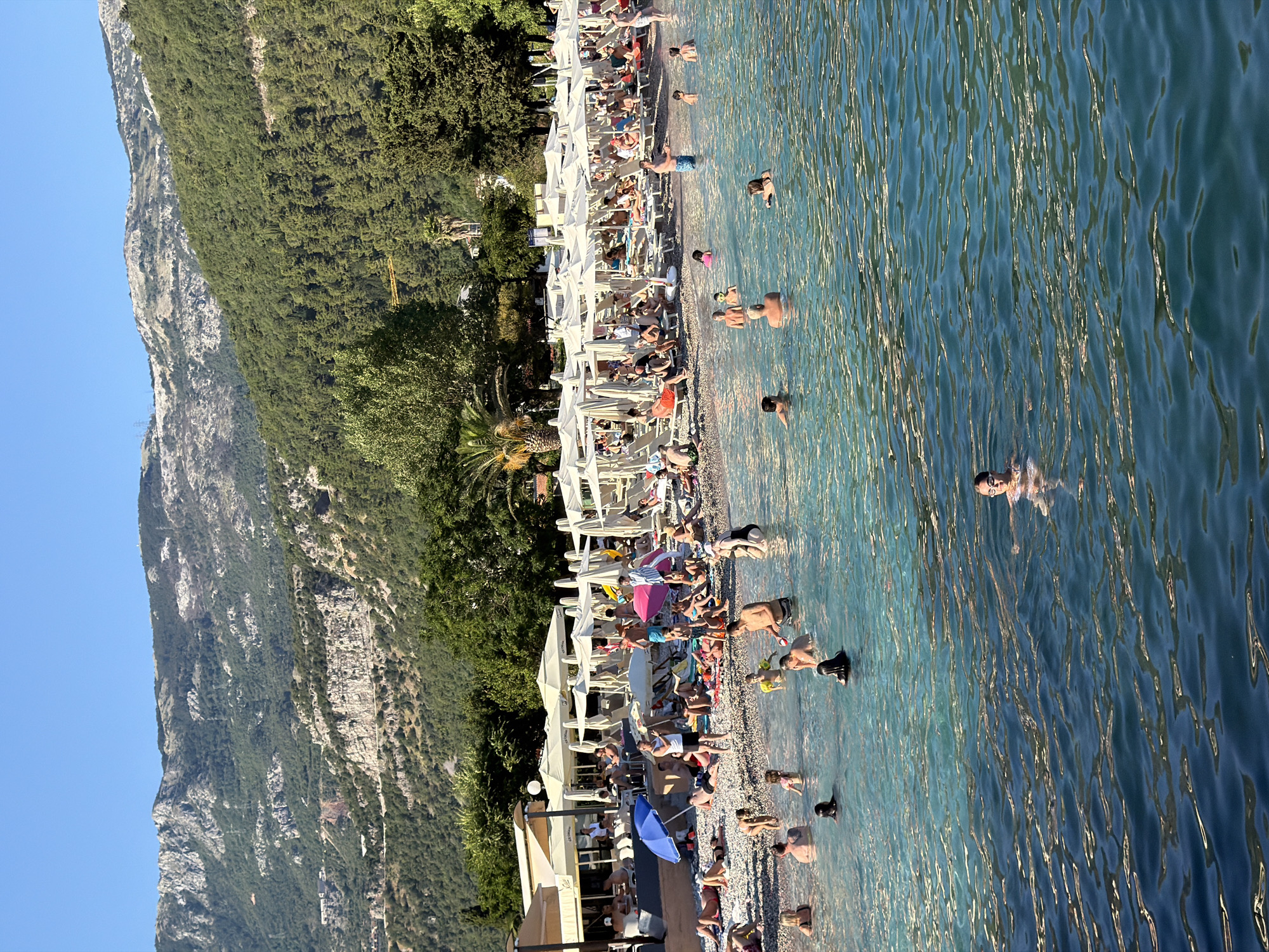The image depicts a sunny day at a crowded beach in Budva, Montenegro. The foreground is dominated by the clear, blue-green water of the Adriatic Sea, with several people swimming and wading. A young girl in goggles is visible on the right, partially submerged. Further back, the pebbly beach is lined with numerous white umbrellas and lounge chairs, occupied by sunbathers. Behind the beach, lush green trees and vegetation climb up the steep, rocky mountains that form the backdrop of the scene. The sky is a clear, bright blue.
