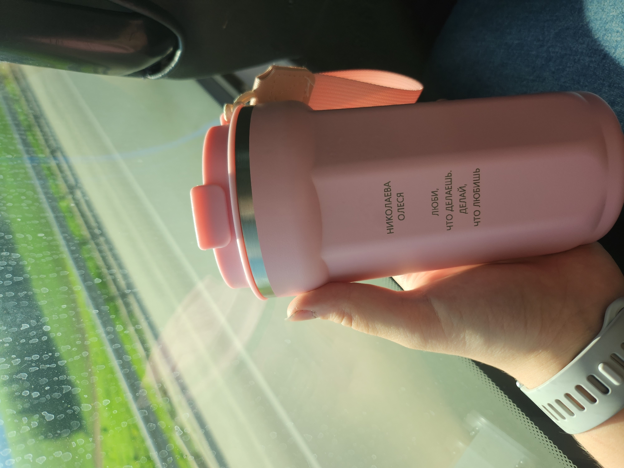 A person is holding a pink thermos in their hand. The thermos has the inscription "НИКОЛАЕВА ОЛЕСЯ" and "ЛЮБИ, ЧТО ДЕЛАЕШЬ, ДЕЛАЙ, ЧТО ЛЮБИШЬ". The person is wearing a gray smartwatch on their wrist. The background shows a car windshield with a view of trees and grass. The location is a car in an unknown city.