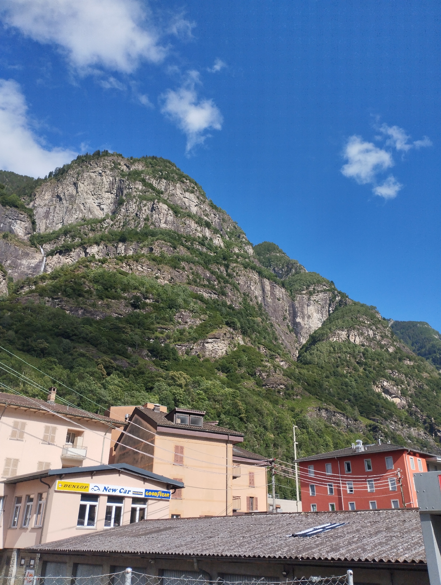 The image captures a view of a town nestled at the base of a steep, rocky mountain. The mountain is covered with lush green vegetation, and a thin waterfall cascades down its side. In the foreground, there are several buildings, including a peach-colored building with a sign that reads "Dunlop New Car Goodyear". To its right is a vibrant red building with numerous windows. The sky above is a clear blue with scattered white clouds. The scene appears to be a sunny day in a mountainous region.
