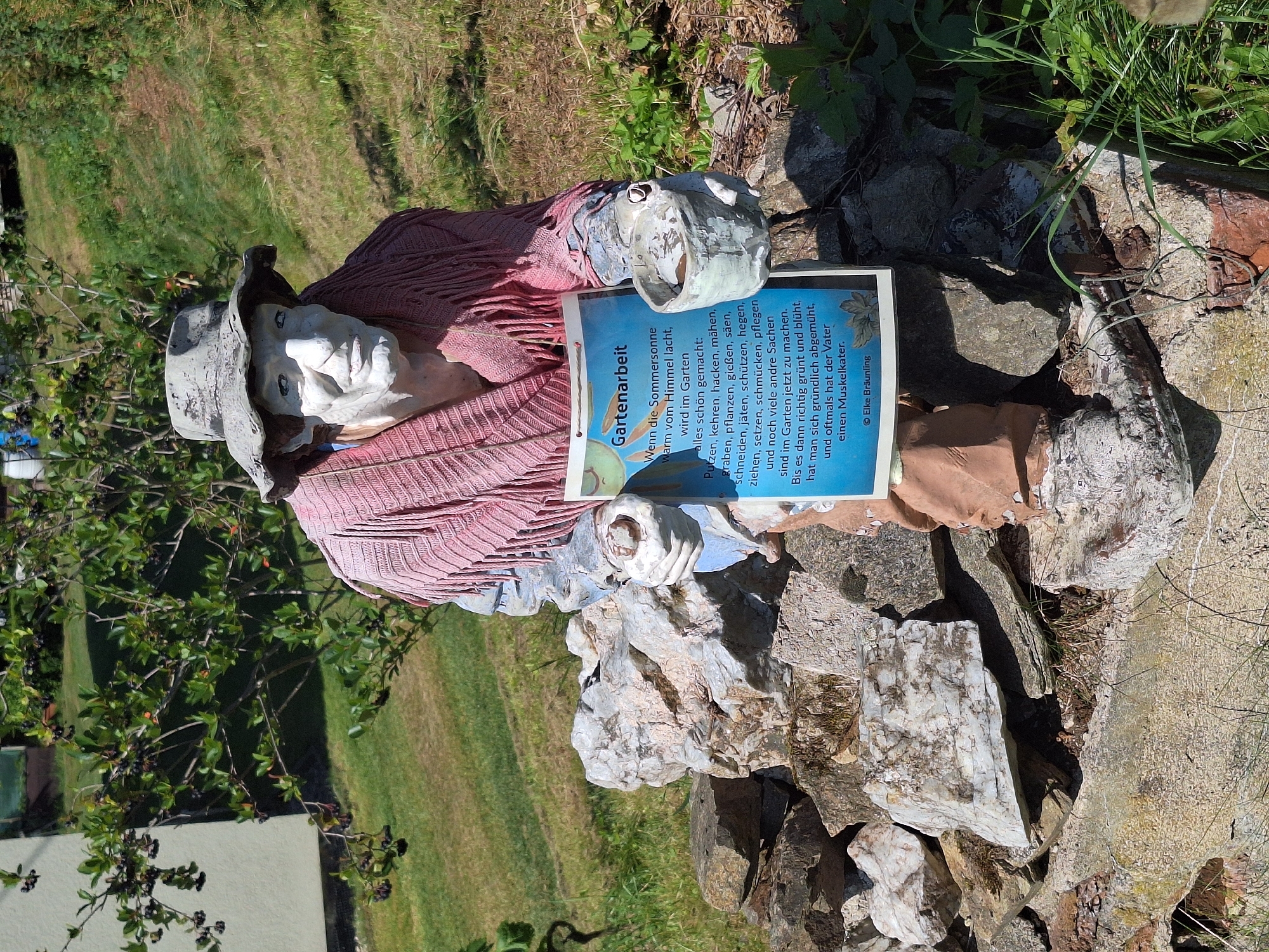 A full-body, eye-level, slightly low-angle shot captures a life-sized, white-faced figure, dressed as a gardener, standing amidst a pile of rough-hewn stones and patchy green grass. The figure wears a wide-brimmed, weathered grey hat and a pink, fringed shawl draped over its shoulders. Its body appears to be constructed from concrete or plaster, with visible brushstrokes and a rough texture. The figure's face is stark white, with hollowed eyes, a prominent nose, and a faint smile. It holds a light blue, rectangular sign in its left hand, featuring text in German and a small illustration of a plant. The sign is titled "Gartenarbeit" (Gardening). In its right hand, the figure holds a small, grey, bucket-like object. The background consists of a gently sloping, grassy hillside with scattered rocks and some green foliage, including a bush with dark berries to the left of the figure. In the far upper left, a portion of a light-colored building is visible. The scene is brightly lit by sunlight, casting shadows on the ground and the figure. The overall impression is that of an outdoor garden decoration or a whimsical artistic representation of a gardener at work. The provided text on the sign refers to gardening activities and the arrival of summer, and is attributed to "© Elke Bräunling". This imagery is consistent with garden art often found in rural or semi-rural settings.
