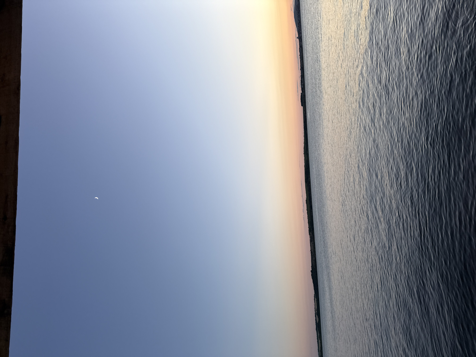 The image depicts a serene twilight scene over a body of water, likely the Burgas Bay in Burgas, Bulgaria. A sliver of a crescent moon is visible in the clear, pale blue sky, which transitions to soft hues of orange and pink near the horizon. The water's surface is rippled, reflecting the subtle colors of the sunset. In the distance, a dark, elongated landmass, presumably the Burgas coast, is visible with a faint treeline. The foreground is dominated by the dark, textured surface of the water, creating a sense of depth. A dark, wooden structure, possibly part of a pier or balcony, juts into the frame from the top edge, providing a sense of vantage point. There are no people, animals, or specific identifiable structures on the distant shore visible in detail.