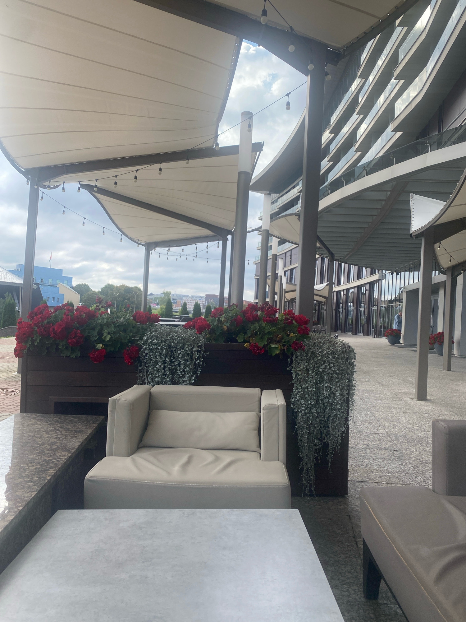 The image captures an outdoor seating area of a modern building complex in Minsk, Belarus. In the foreground, a light-colored sofa and a matching armchair are placed next to a textured, light gray coffee table. Beyond the seating, a planter box filled with vibrant red flowers and trailing green plants is visible. The seating area is covered by a white awning supported by dark pillars, with string lights illuminating the space.
In the background, a multi-story building with a contemporary facade featuring large windows and balconies curves into the distance. The ground is paved with speckled gray stones. The sky is overcast with scattered clouds. The overall scene suggests a well-maintained, urban outdoor dining or lounge area.