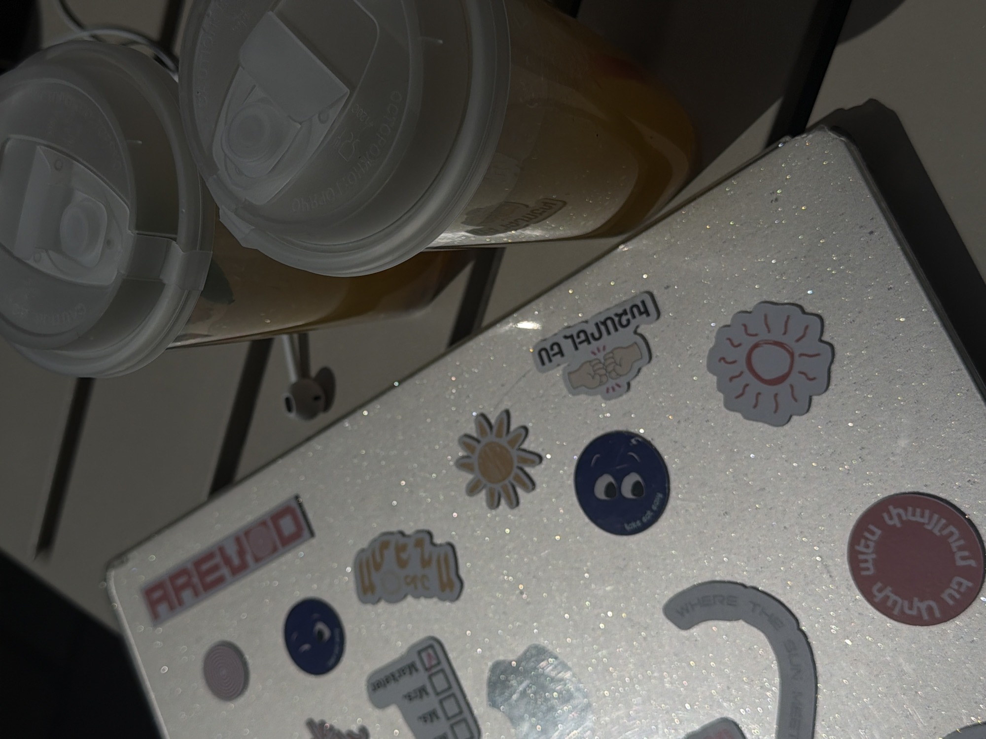 The image is a close-up shot taken from an overhead perspective, featuring two sealed cups filled with a translucent, yellowish liquid, likely a beverage. The cups are positioned to the left of a square, flat surface, which is adorned with a series of colorful stickers. The stickers include various designs such as suns, faces, text in Armenian and English, and check-box icons, suggesting a customized or personalized item. The flat surface has a sparkling, glittery finish. In the background, hints of a white, slatted surface are visible.