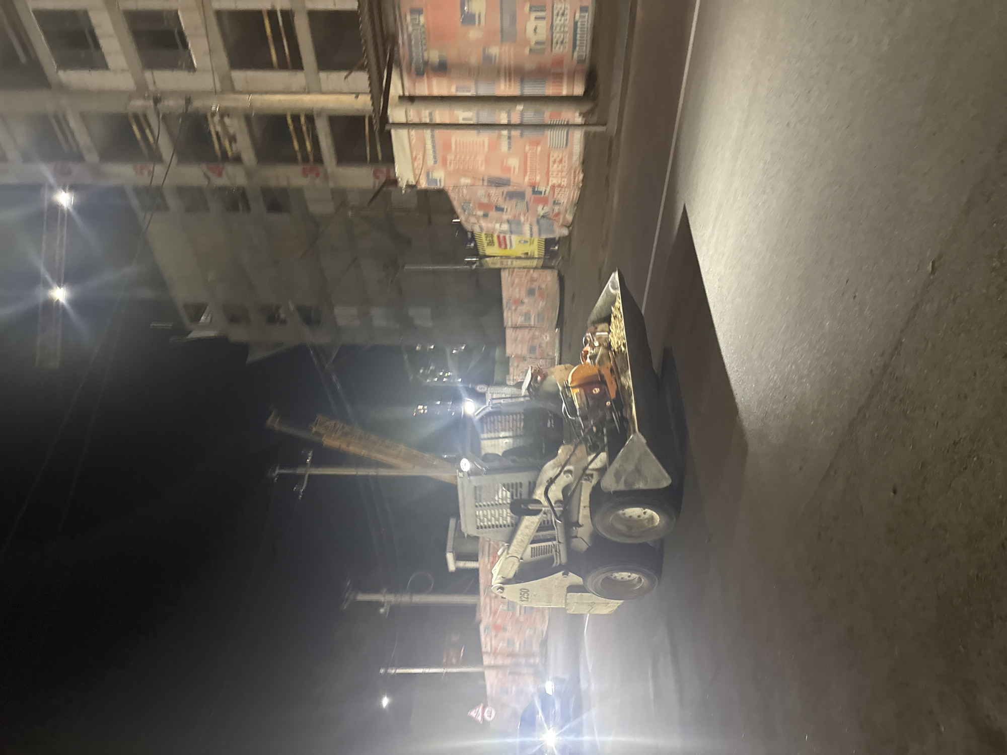 In Almaty, Kazakhstan, at night, a small excavator with a front loader is parked on a paved road, illuminated by artificial lights. The excavator's bucket contains a pile of gravel and what appears to be a circular saw. In the background, a multi-story building under construction is visible, with scaffolding and construction materials. The building is covered with protective netting adorned with colorful advertisements or images. A street light illuminates the road in front of the excavator, casting a bright beam. Power lines are strung overhead, and a traffic sign is visible on the left side of the image. The scene suggests ongoing construction work at night.