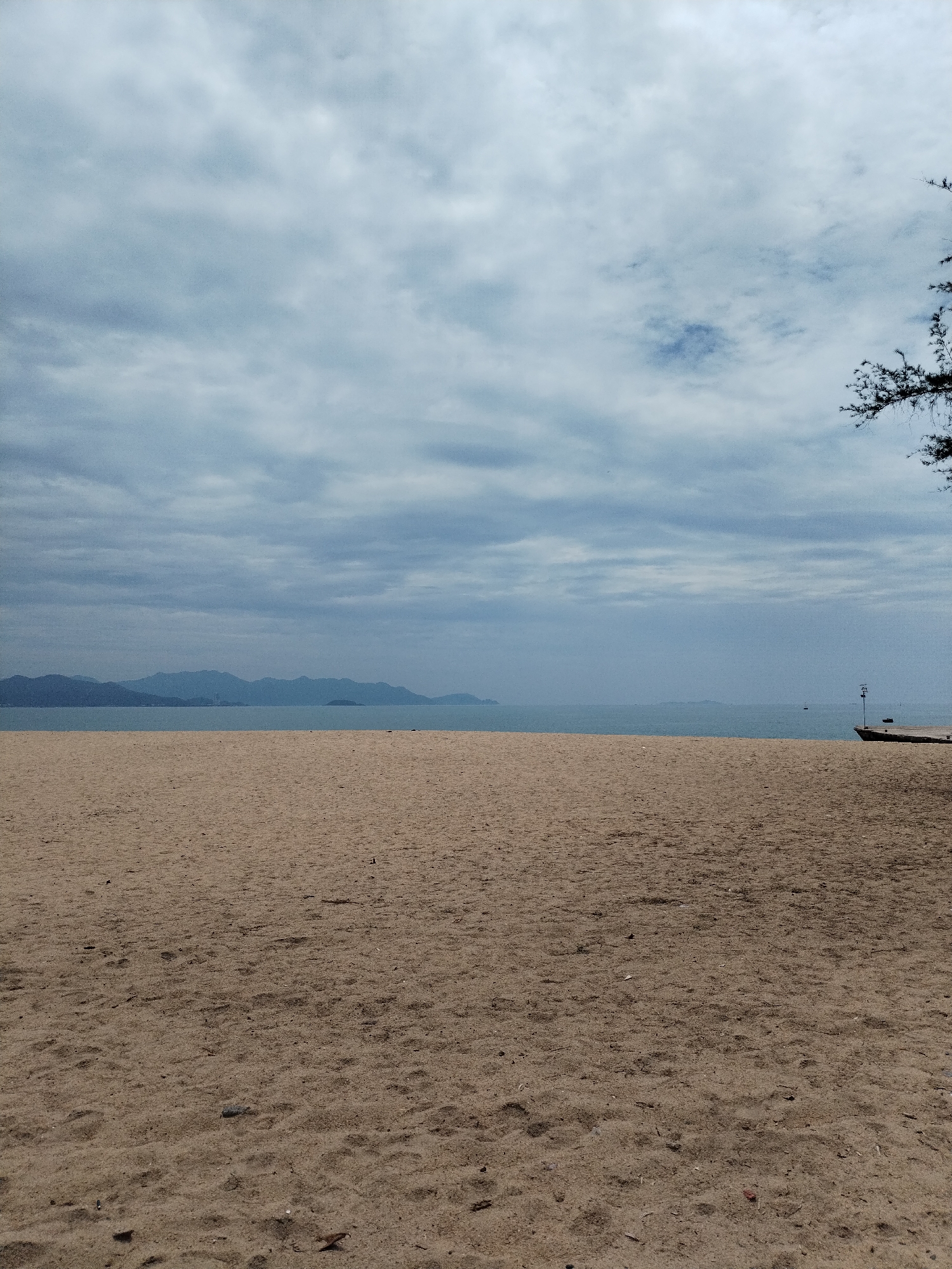 This media file presents a wide-angle landscape view of an overcast beach scene in Nha Trang Ward, Vietnam. The image is divided into three main horizontal sections: a sandy beach in the foreground, the sea in the midground, and a cloudy sky with distant mountains in the background.
The extensive sandy beach occupies the lower half of the frame. The sand is light brown with varying textures, showing some subtle undulations and scattered small pieces of debris like twigs and leaves. There are no clear footprints or other human traces visible on the immediate sand, suggesting an undisturbed area.
Beyond the beach, the calm, grey-blue sea stretches towards the horizon. In the far distance, a low chain of mountains or hills, appearing hazy and dark blue-grey, rises from the water, spanning much of the left and central horizon. The sky above is entirely covered by a dense layer of diffused, medium-grey clouds, indicating an overcast day with no direct sunlight.
On the far right side of the image, the dark green foliage of a tree or large bush is partially visible, extending into the frame from the top right corner. Closer to the waterline on the right, a dark, indistinct structure or object, possibly the bow of a small boat or a small wooden pier, can be seen with a slender pole extending upwards.
There are no people visible, and no specific activities are taking place. The overall mood is one of quietude and tranquility, characteristic of a calm, overcast day at the beach. No visible text is present in the image.