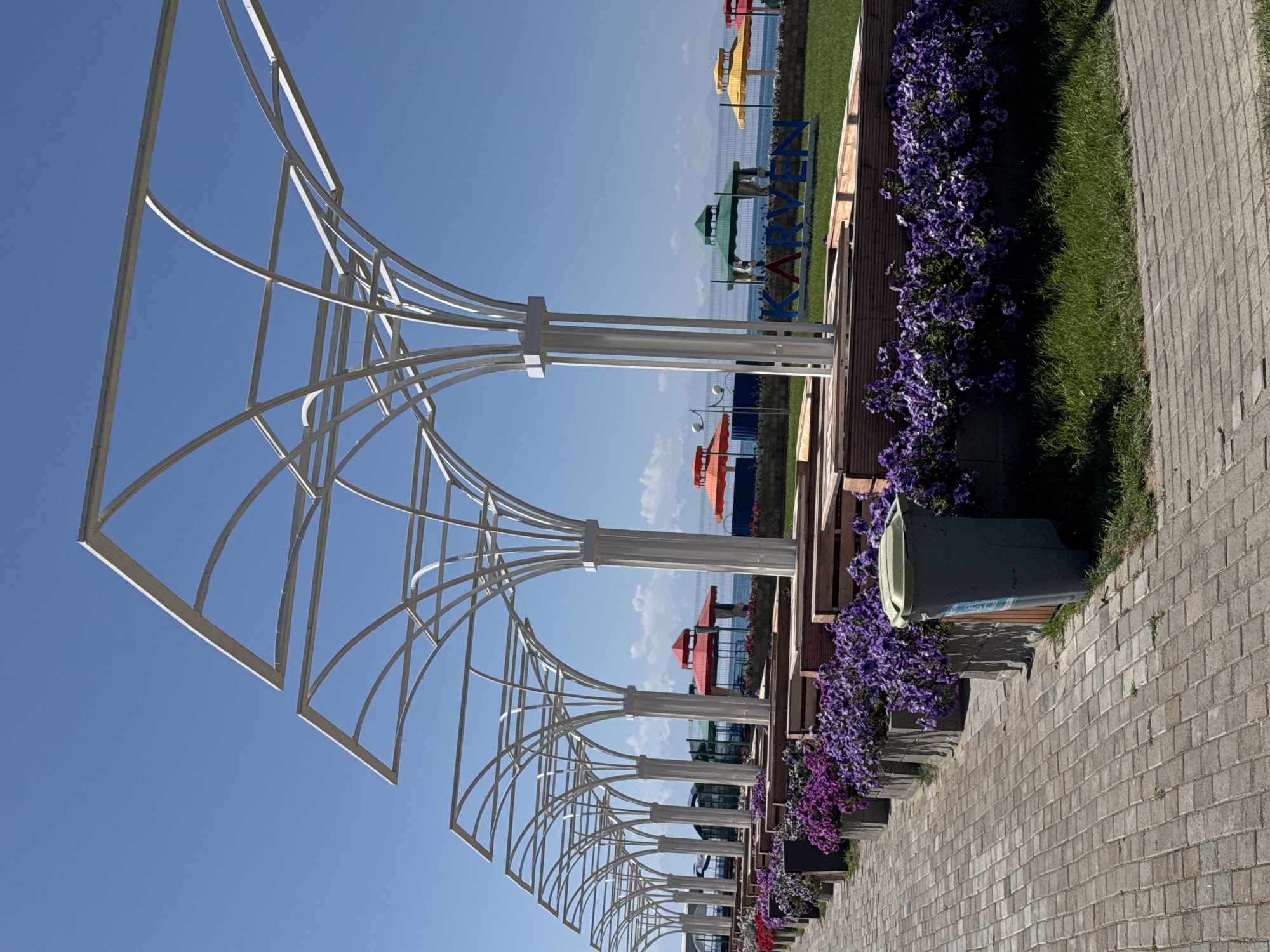 In Sary-Oy, Kyrgyzstan, a paved walkway runs alongside a row of planters filled with vibrant purple petunias. Beyond the planters, wooden benches are arranged, providing seating. A series of white, tree-like canopy structures with curving metal supports stretch along the walkway, offering shade. In the background, a grassy area features a sign with the letters "KARVEN" in blue, red, and green. Beyond the grass, a body of water is visible, with several colorful gazebos and umbrellas, including red, green, yellow, and orange, scattered along the shoreline. Mountains are visible in the distance, under a clear blue sky. A grey trash can stands on the paved walkway near the planters. The scene appears to be a recreational area near a lake.