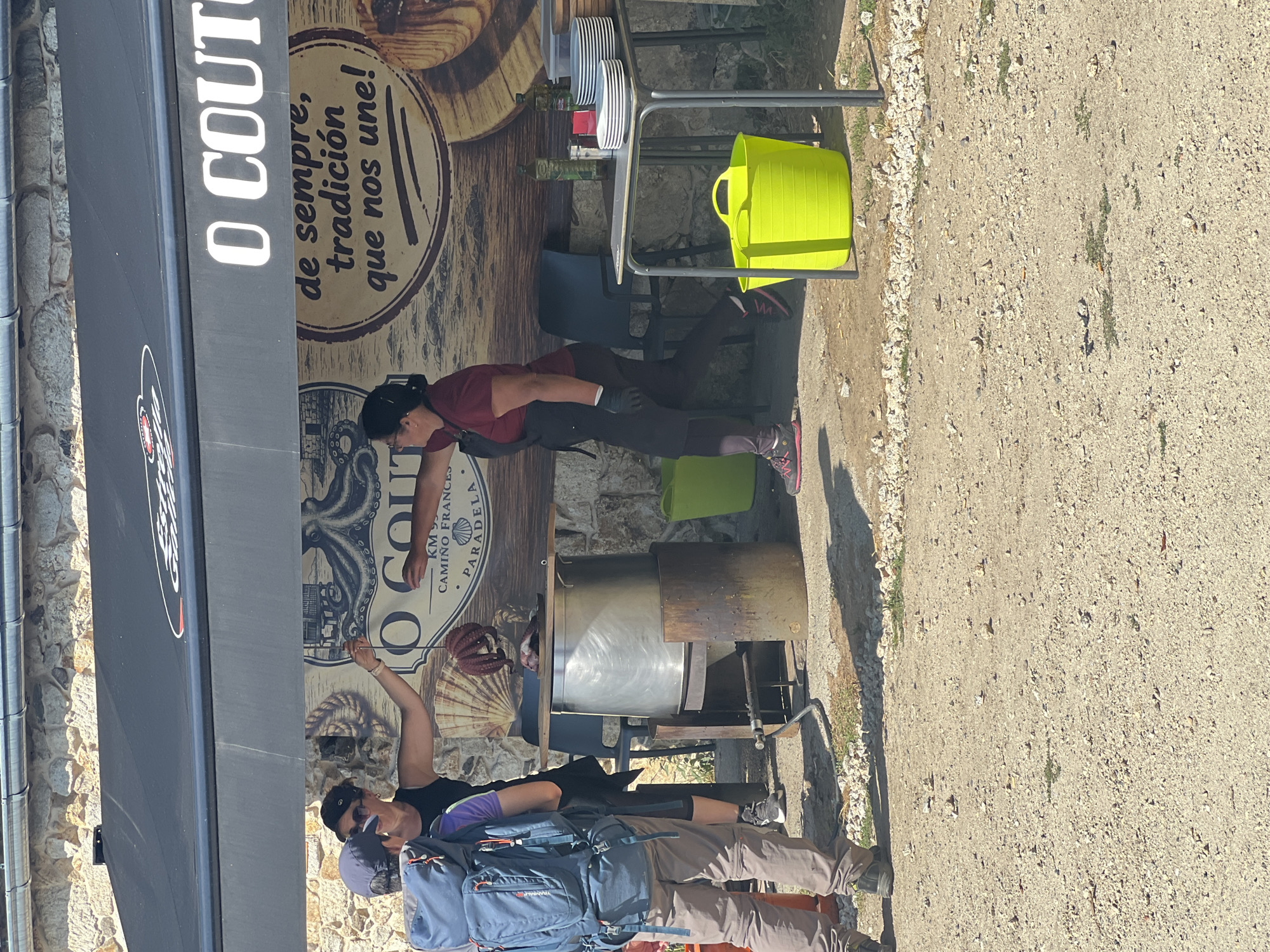 In Paradela, Spain, a street-side establishment displays food and beverages for sale, possibly related to a local festival or market. A large metal pot, heated by a portable stove, is prominent in the scene, suggesting food preparation. Several individuals are present, engaged in various activities. One person, wearing a backpack, stands on the left side of the image. Another person, dressed in an apron and gloves, appears to be serving or preparing food. Behind them, a woman in dark clothing and a cap holds a string with an octopus and some meat suspended from it. A table to the right is laden with plates and bottles, indicating a serving area. The environment is outdoors, with a stone wall and a black awning providing shelter. The ground is covered with gravel and dirt. The overall atmosphere suggests a bustling commercial setting with a focus on food service.