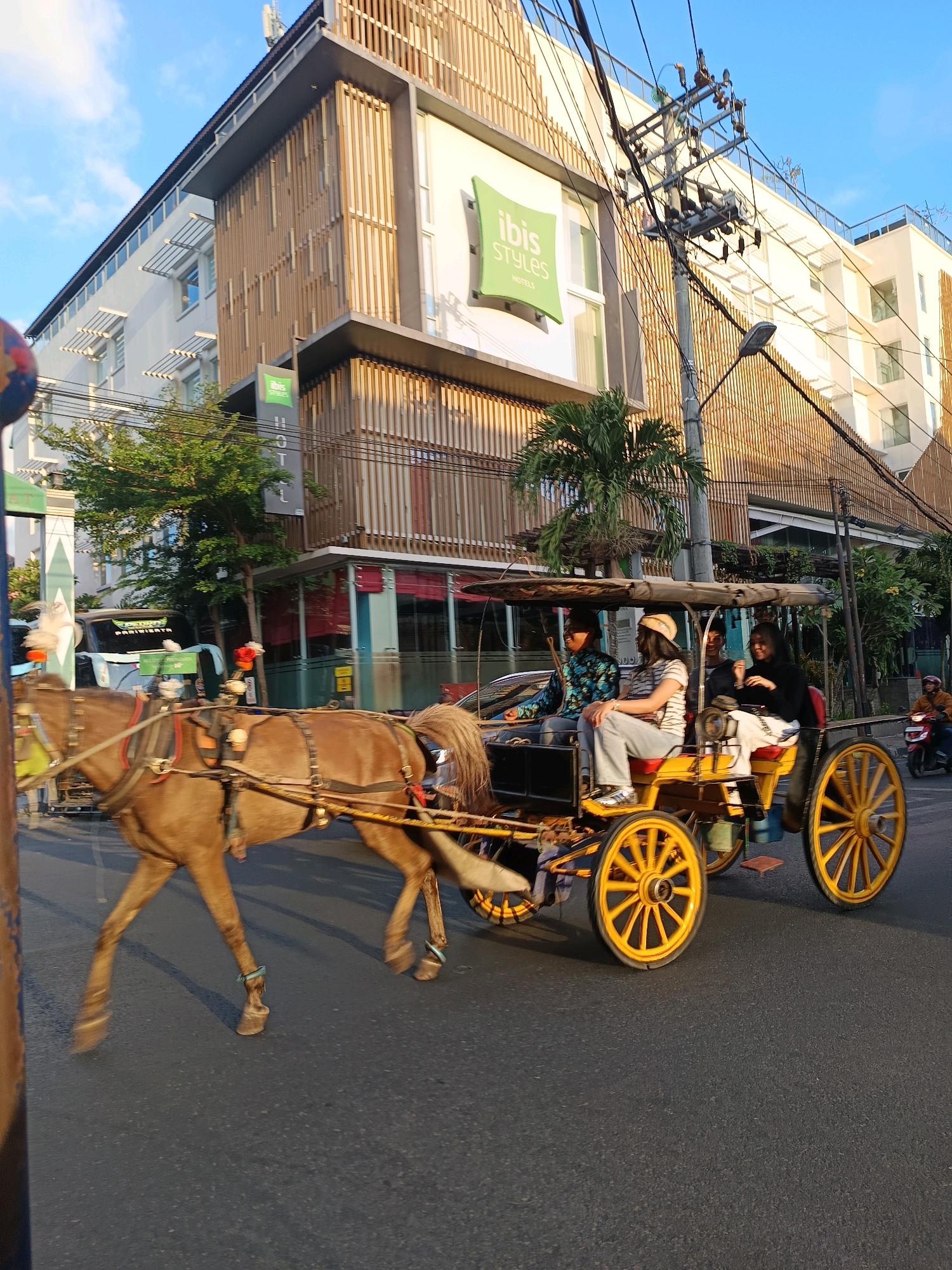 A horse-drawn carriage, known locally as an Andong, travels down a street in Yogyakarta, Indonesia. The carriage is yellow with bright yellow spoked wheels and is pulled by a brown horse. Three people are seated in the carriage; two women and one man. The man is driving, and the women are looking in different directions. The carriage is moving on the right side of the road, passing a white building with wooden accents. The building has a sign that reads "ibis STYLES HOTELS". Overhead, there are power lines and a lamppost. The sky is blue with some clouds. The street is paved with asphalt, and there are trees and other vegetation visible. In the background, a person on a motorcycle is also traveling down the street. The scene appears to be in an urban environment.