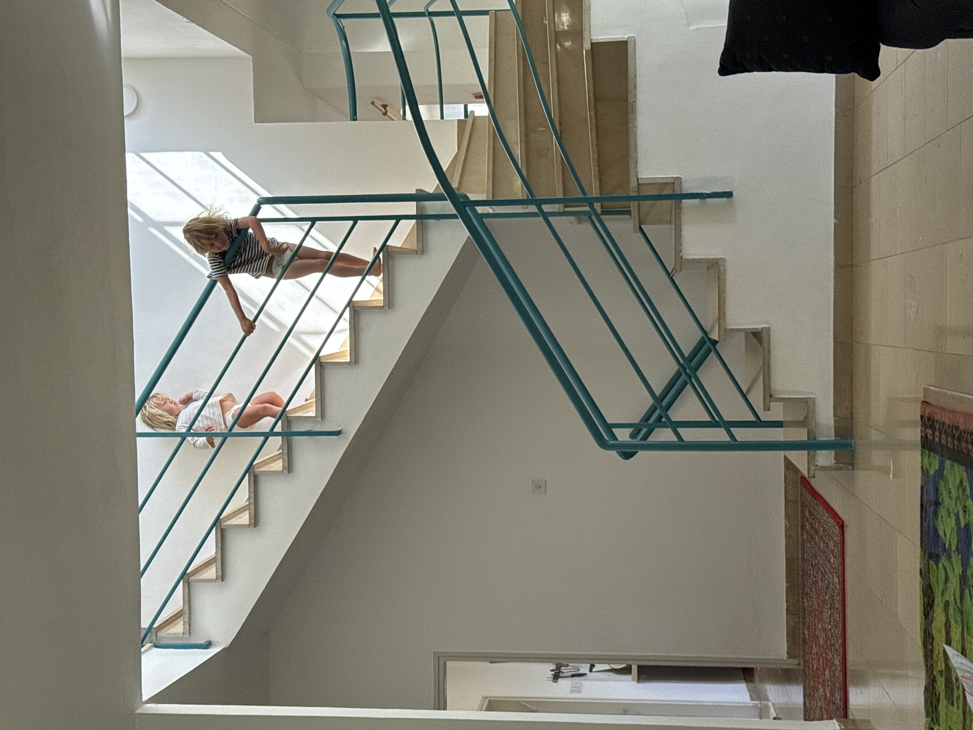 An interior scene in Kfar Vradim, Israel, depicts two young children on a multi-level staircase with a teal metal railing.
The environment consists of white walls, light tan/cream tiled flooring, and matching marble-effect stair treads with white risers. The prominent railing system is constructed from teal-colored metal, featuring a top handrail and multiple horizontal bars.
On the upper section of the staircase, a younger child with light blonde hair, wearing a white long-sleeved top and white underwear/diaper, stands facing right, holding the teal railing with both hands. Below and to the right of this child, a slightly older child with light blonde hair, wearing a striped short-sleeved top and white underwear/diaper, stands on a lower step, leaning forward and holding the railing with their left hand. Both children appear to be observing something below them or engaging with the stairs.
Strong natural light enters from an unseen source above and to the right, casting distinct diagonal shadows of the railing onto the white wall behind the children.
In the left foreground, a red patterned rug is partially visible on the tiled floor, leading to an open doorway revealing a glimpse of an interior hallway with objects on a wall fixture and a shelf. Another multicolored rug is partially visible in the bottom-left corner. On the white wall to the left of the staircase, an electrical outlet or switch is visible. A dark pillow is partially visible in the bottom-right foreground.