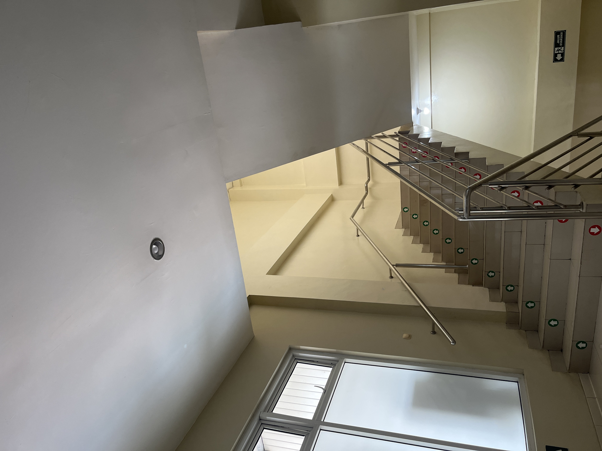 The image shows an interior view of a staircase with a large window on the left. The staircase is made of light-colored tiles and has a stainless steel railing. Green arrow stickers indicating upward movement are placed on the steps. Red arrow stickers, presumably indicating downward movement or an alternate path, are also visible on some steps. A sign with the text "JALUR EVAKUASI" and an evacuation symbol is affixed to the wall on the right side of the image, indicating an emergency exit route. The walls are painted in a light yellow hue, and the ceiling is white with a recessed light fixture. The environment appears to be a public building or a common area within a building. The location is identified as Kedungdowo, Indonesia.