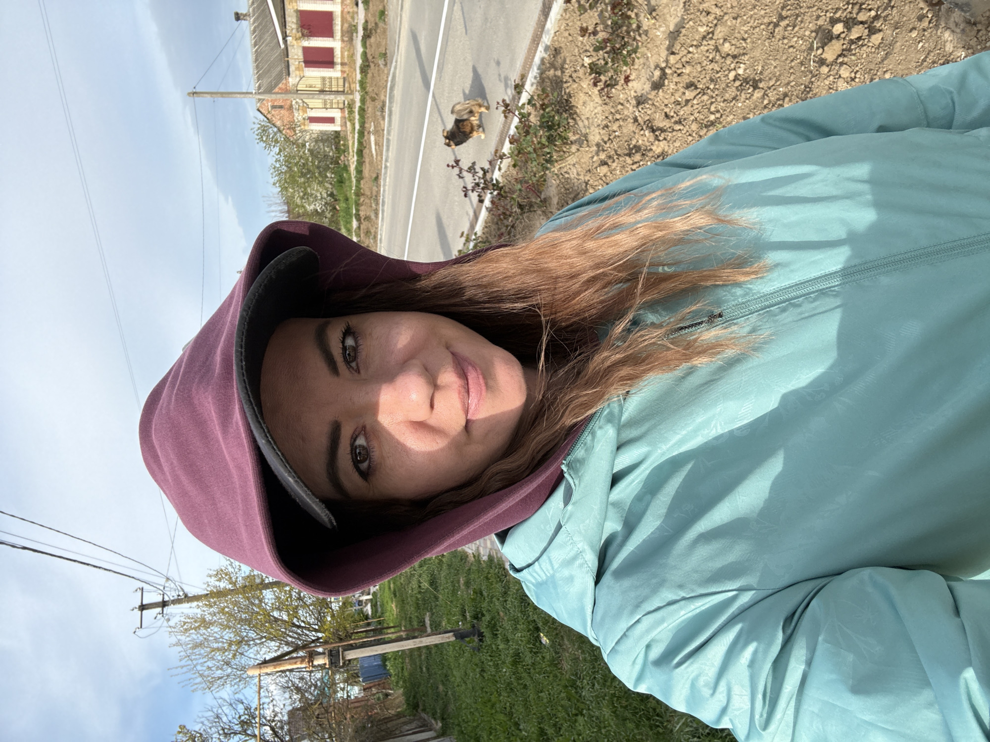 A woman with auburn hair wearing a light blue jacket with a maroon-lined hood stands outdoors on the edge of a road in Taman, Russia. A small dog stands on the edge of the road next to a building with red window frames. Telephone poles run along the roadside and through the landscape behind the woman.