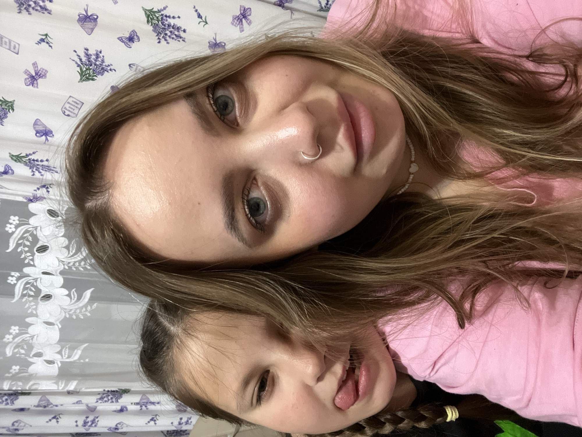 This image features two females, an adult and a child, captured in a close-up, informal portrait. The photograph appears to be taken from a top-down perspective and is rotated roughly 90 degrees counter-clockwise.
The adult, positioned towards the top of the frame, has long, wavy light brown hair with blonde highlights and blue-grey eyes. She wears a small silver hoop nose ring and a white beaded necklace adorned with a heart charm. Her expression is calm and neutral. She is wearing a pink garment.
The child is visible below the adult, partly obscured by the adult's hair. She has light brown hair, with a braid secured by a yellow elastic band visible on the right side of the frame. She is playfully sticking out her tongue, indicating a lighthearted and jovial mood. The child also appears to be wearing a pink top.
The background consists of patterned fabric, likely bedding or upholstery, suggesting an indoor setting. One dominant pattern on a white base features recurring motifs of purple lavender sprigs, bows, butterflies, and small, illegible textual shapes. Another section, visible towards the bottom left, shows a grey and white floral design.
No explicit text is discernible within the image. The time of day is indeterminate, and the subjects' emotions range from calm to playful. While the location context is Novopokrovskaya, Russia, there are no specific visual cues within the image to identify this geographical location.