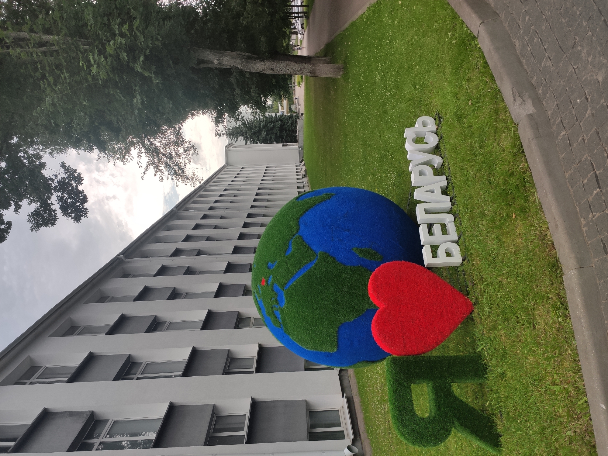 The image depicts an outdoor scene in front of a modern, grey and white building in Navapolatsk, Belarus. In the foreground, there is a large, spherical globe covered in green artificial grass, with blue areas representing oceans and a red area indicating a location on the European continent, likely Belarus itself. To the left of the globe are two large, green, grassy letters forming the Cyrillic letter "Я" (Ya, meaning "I"). Next to "Я" is a large, red, heart-shaped object, also covered in what appears to be artificial grass or a similar textured material. To the right of the heart are white, three-dimensional letters spelling "БЕЛАРУСЬ" (Belarus). The ground is covered in green grass, with some small white wildflowers. The scene is bordered by a paved walkway and a grey concrete curb. Behind the globe and letters, a section of the building is visible, characterized by repetitive windows and dark grey architectural elements. To the right of the building, tall trees and a row of parked cars line a street. The sky is overcast with clouds. A few people are visible in the distance walking along the street and near the cars.