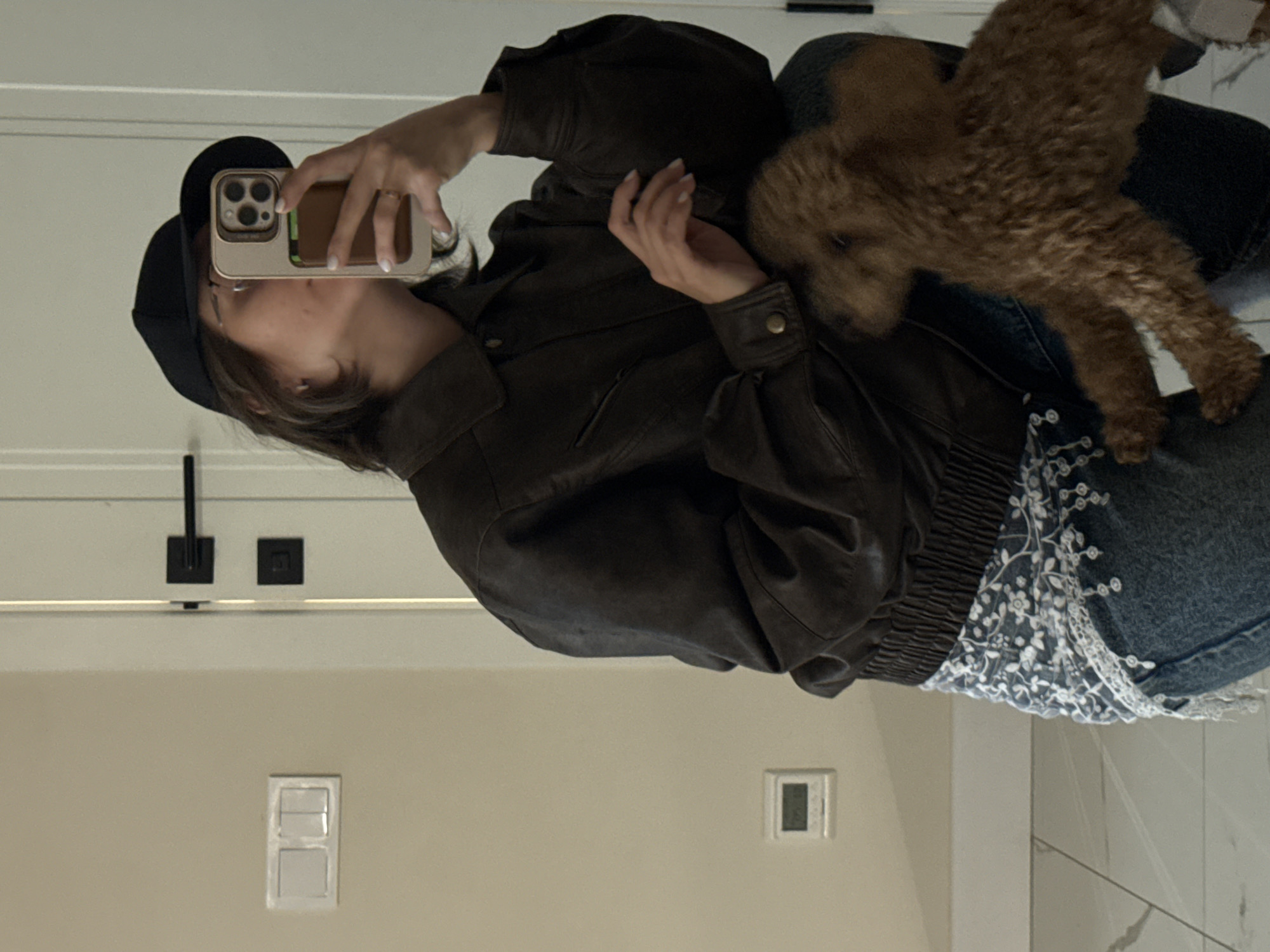 The image, originally rotated 90 degrees counter-clockwise, depicts a person taking a mirror selfie indoors with a small dog. The main subject, appearing to be a young woman, is visible from the chest up, wearing a black baseball cap and a dark brown, possibly leather, bomber jacket with puffed sleeves and an elasticated waistband. She holds a gold or tan-colored smartphone (resembling an iPhone 14 Pro with a triple-lens camera setup) to capture her reflection. A ring is visible on her right ring finger, and her fingernails are painted white.
Perched near her left arm and shoulder is a small, brown, curly-haired dog, looking towards the camera. The person's attire also includes blue jeans featuring a distinctive white lace or embroidered floral pattern on the side, visible beneath the jacket.
The background reveals a plain, light-colored wall or mirror surface, with a white horizontal trim. A black square fixture, possibly a light switch or electrical outlet, is visible, along with another small white rectangular object, likely a light switch, and a tiny square digital display (possibly a thermostat) on the lower wall. The floor appears to be light-toned tiles. The setting suggests a modern indoor environment, such as a home. No specific text is clearly legible, and there are no direct visual cues within the image to indicate the specified location of Pervomaysky, Russia. The lighting is consistent with indoor conditions.