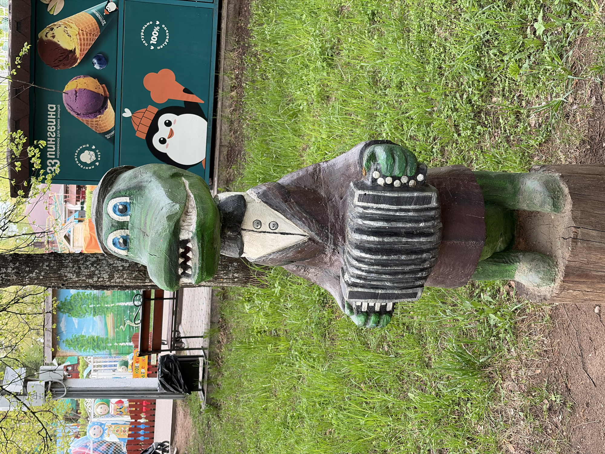 Captured in the Russian city of Chaikovsky, the image features a green wooden sculpture of a crocodile playing an accordion, dressed in a dark brown jacket, white shirt, and small hat. The sculpture is placed on a wooden base amidst a green lawn. To the sculpture's left, an ice cream kiosk features colorful ice cream cones and a penguin drawing, along with the Russian text "33 пингвина" (33 penguins) and "мороженое для ярких эмоций" (ice cream for bright emotions). Further in the background, a painted mural and playground equipment can be observed.