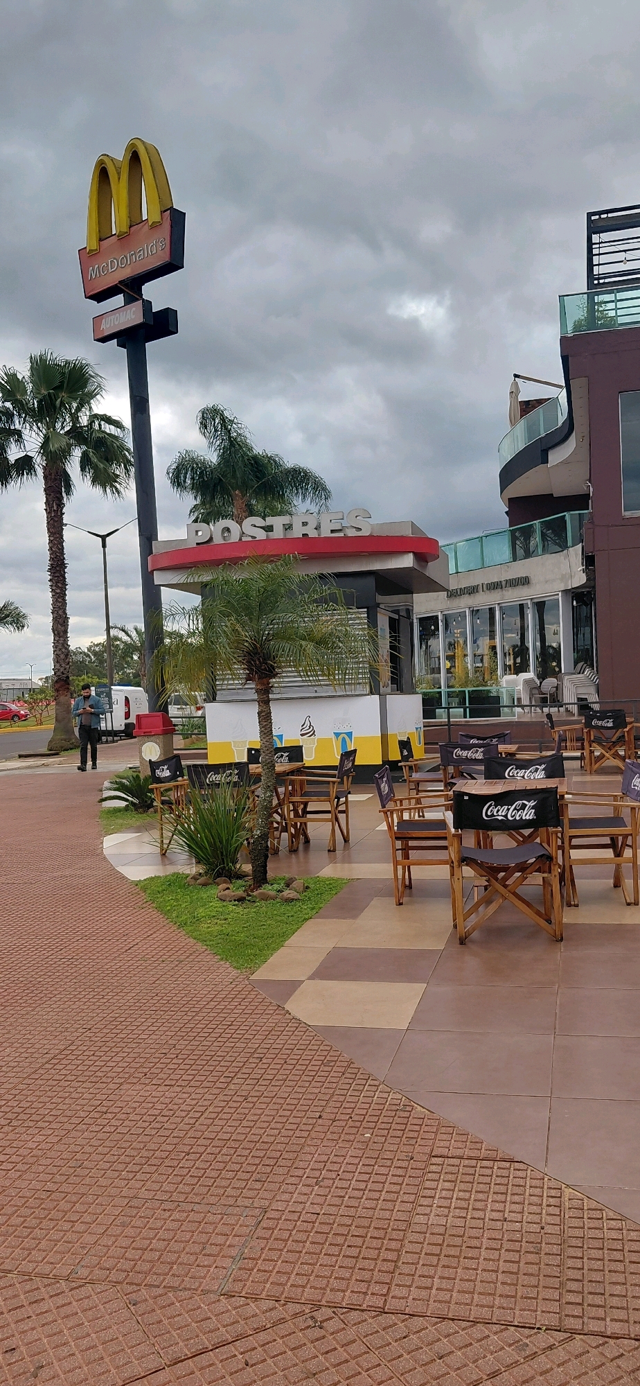 The image shows an outdoor dining area in front of a McDonald's restaurant in Encarnacion, Paraguay. A large McDonald's sign with golden arches and red lettering is visible, along with the "Automac" sign. The "Postres" (Desserts) section of the restaurant is also featured, with a red and white striped awning. Several outdoor tables and chairs with Coca-Cola branding are set up, some with umbrellas. Palm trees and other greenery are present in the scene. A person in a blue shirt is standing near a white van in the background, looking at their phone. The sky is overcast. The ground is paved with reddish-brown textured tiles and some brown and beige square tiles. In the distance, part of a multi-story building with balconies and glass railings is visible.